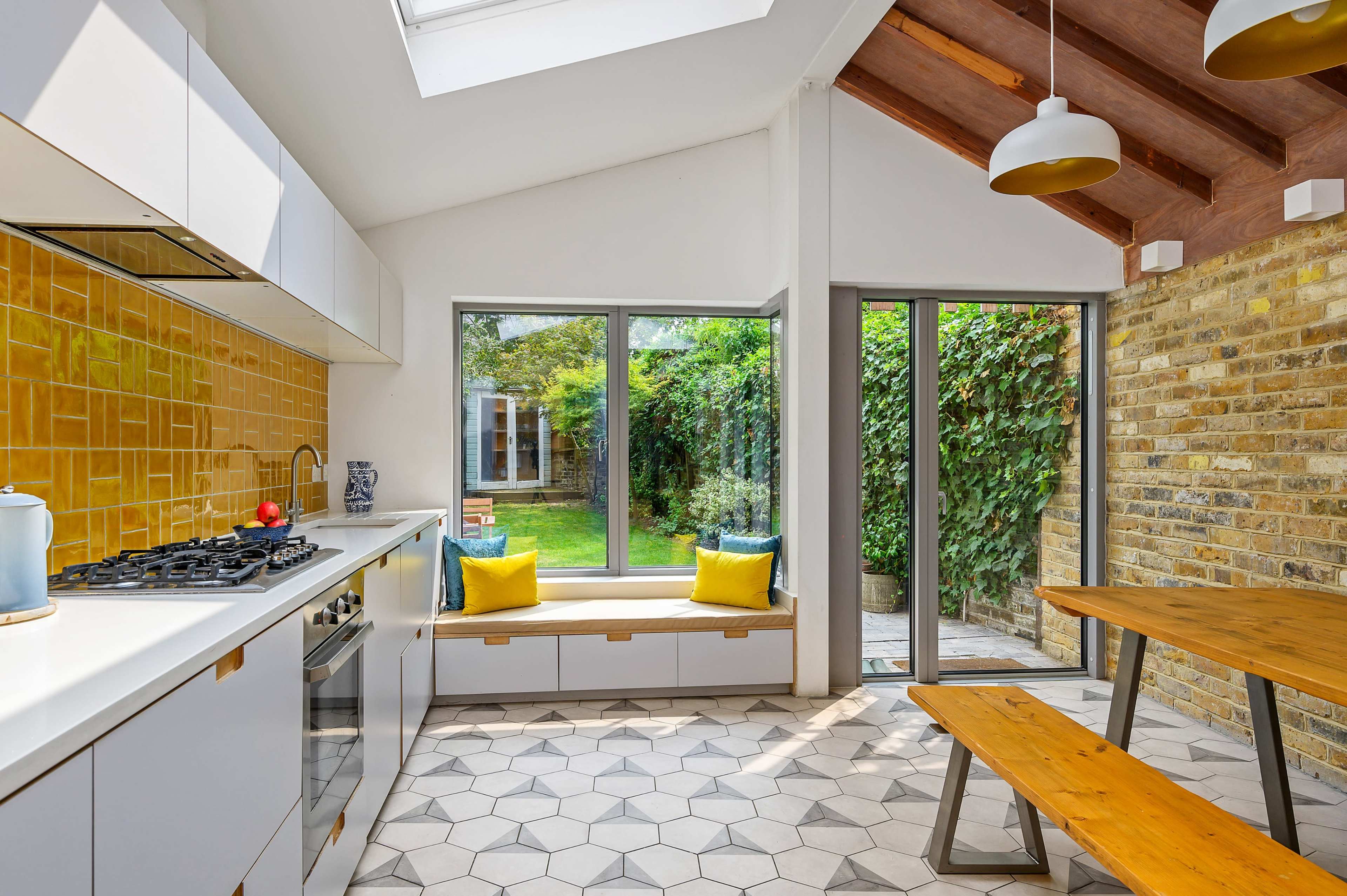 The image shows a modern kitchen with a skylight, featuring white cabinetry, a yellow tiled backsplash, and a dining area with a wooden table and bench seating by large windows overlooking a garden.