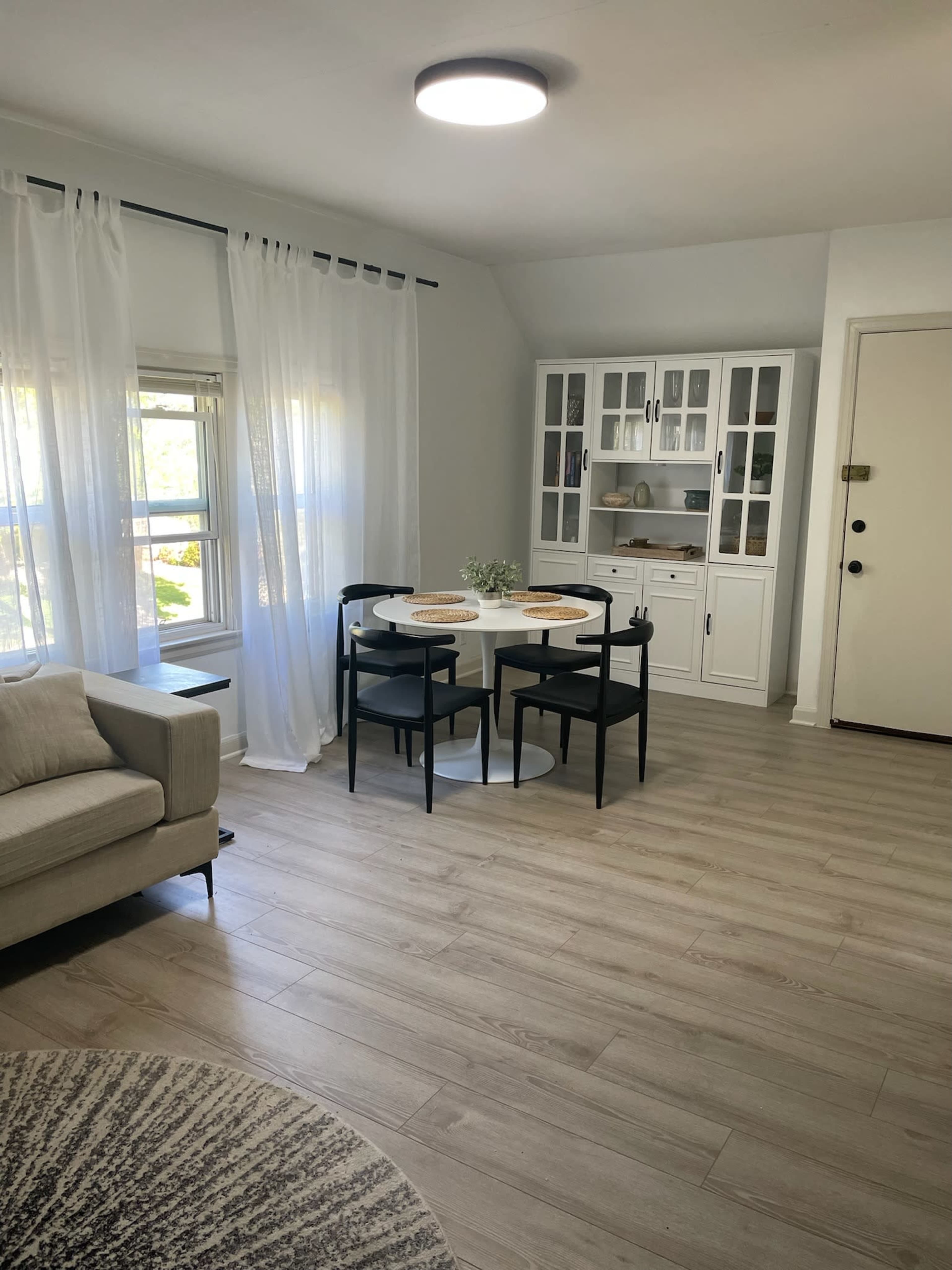 The image shows a bright, modern living space featuring a small dining table with two black chairs, a beige sofa, and a white cabinet against the wall.