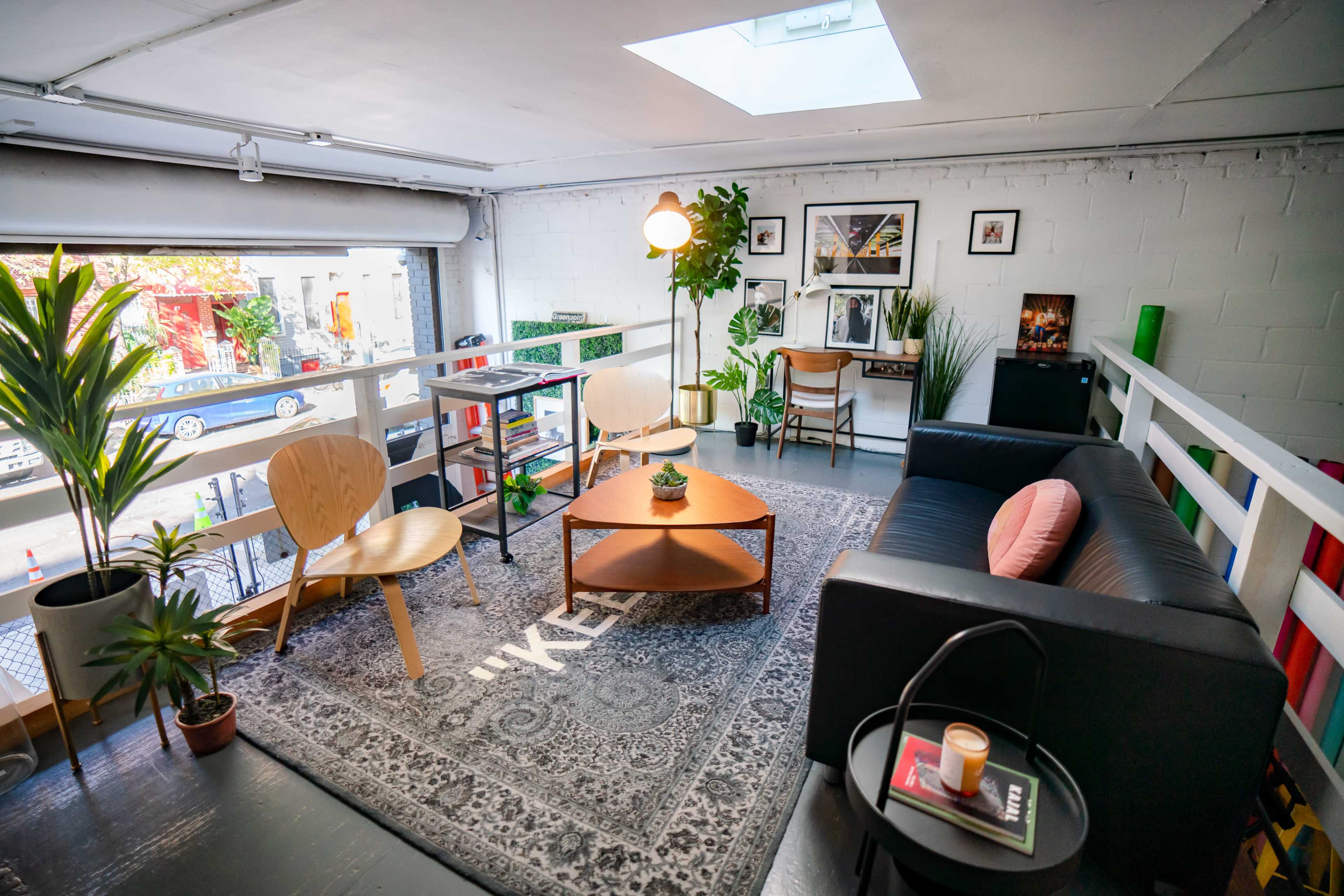 The image shows a modern living space featuring a black sofa, two wooden chairs, a coffee table with a small plant, and various decorative elements, all illuminated by natural light from a skylight.
