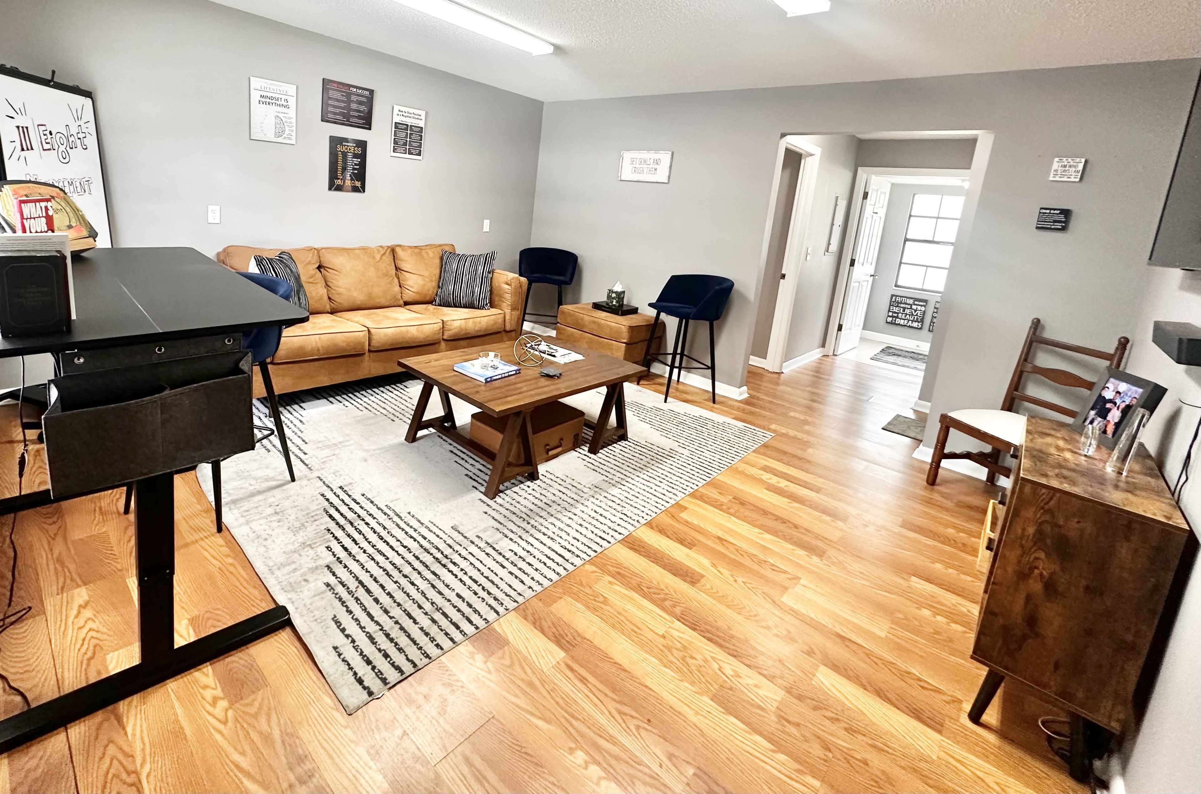 The image shows a modern living room with a brown leather sofa, a wooden coffee table, and hardwood floors, complemented by a rug and seating in a neutral-toned decor.