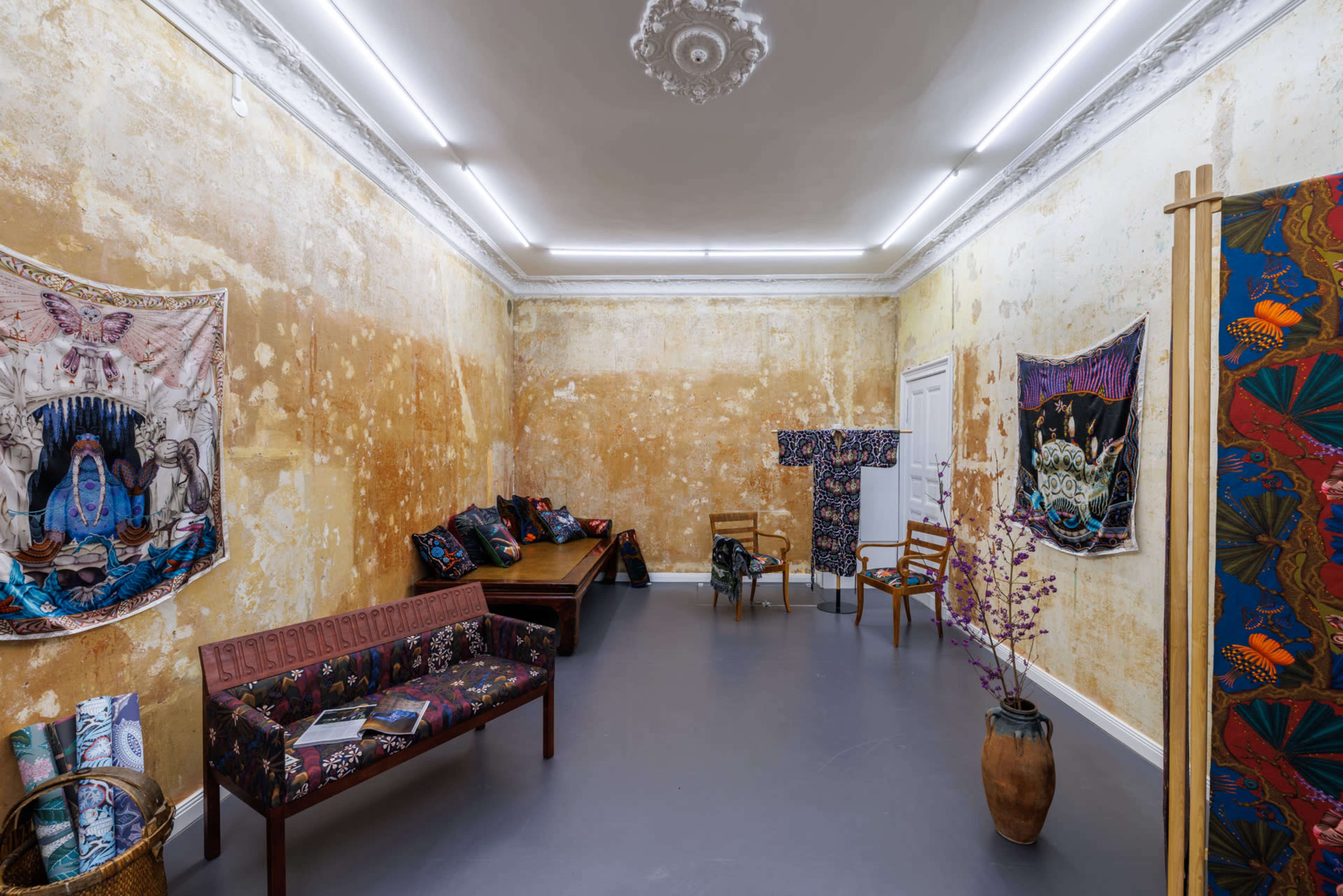 A room with peeling yellow walls, featuring patterned fabric artworks, a wooden bench, chairs, and a decorative vase.