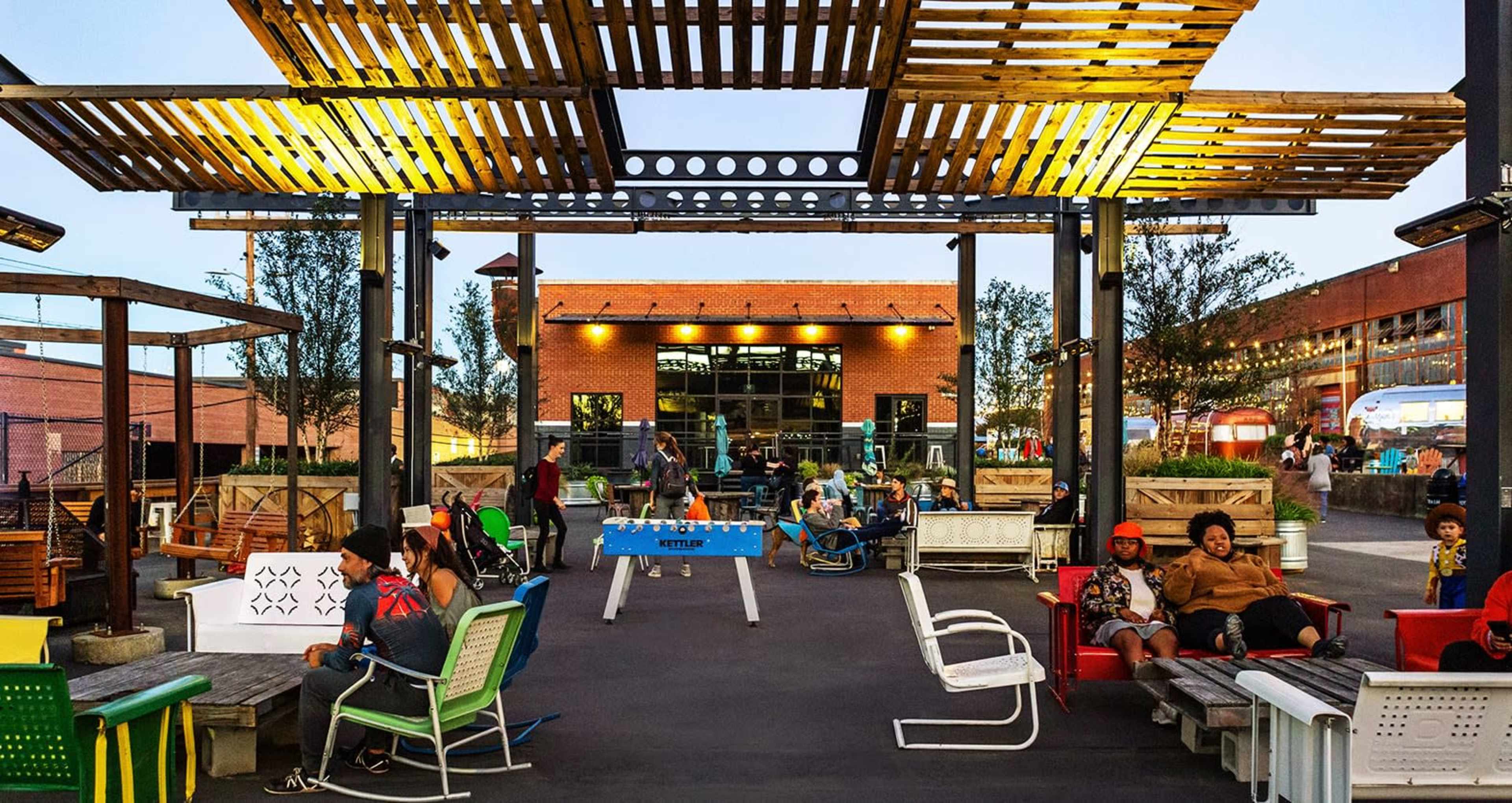 The image shows a lively outdoor area with seating, a table for playing games, and people enjoying the space around a building.