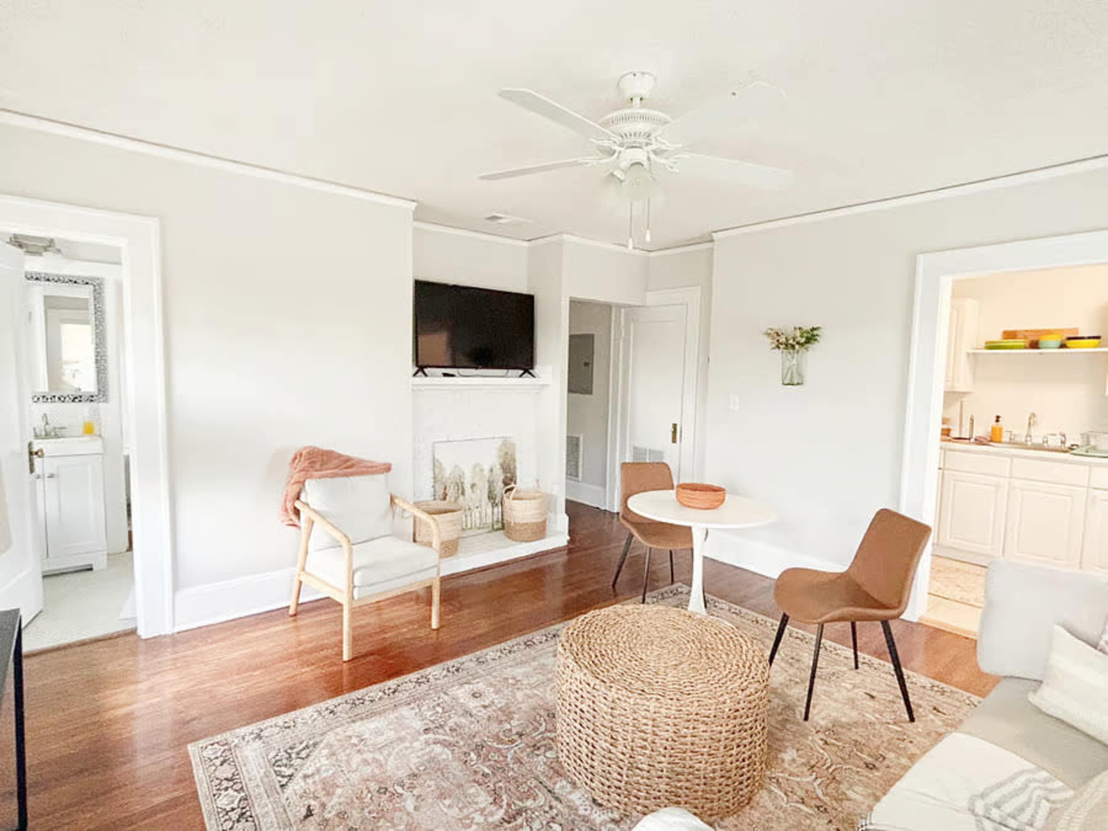 The image shows a living room with a beige color scheme, featuring a fireplace, a television mounted on the wall, a round table, and various seating options.