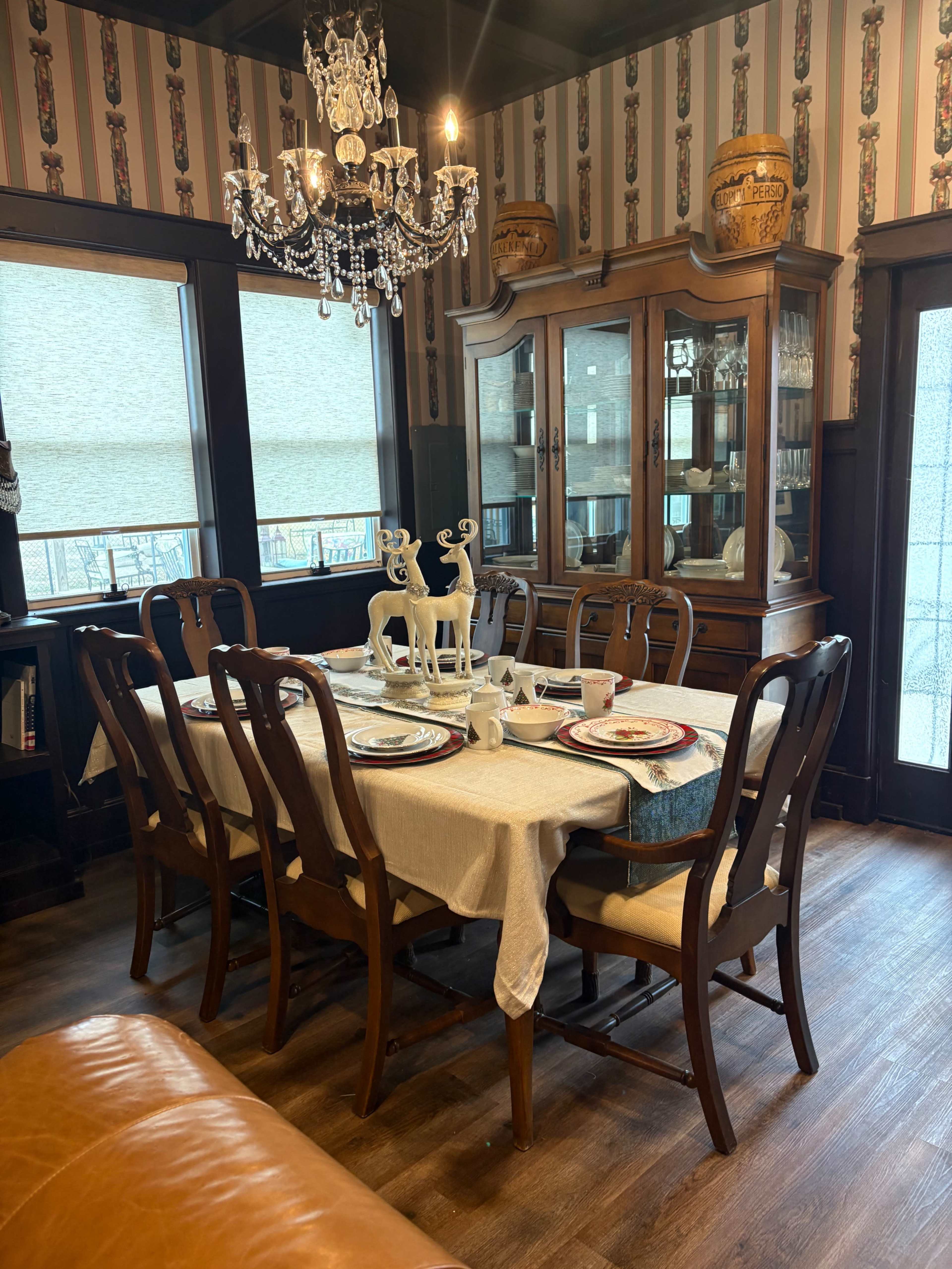 The image shows a dining room featuring a large table set with dishes, surrounded by wooden chairs, and a chandelier hanging above.