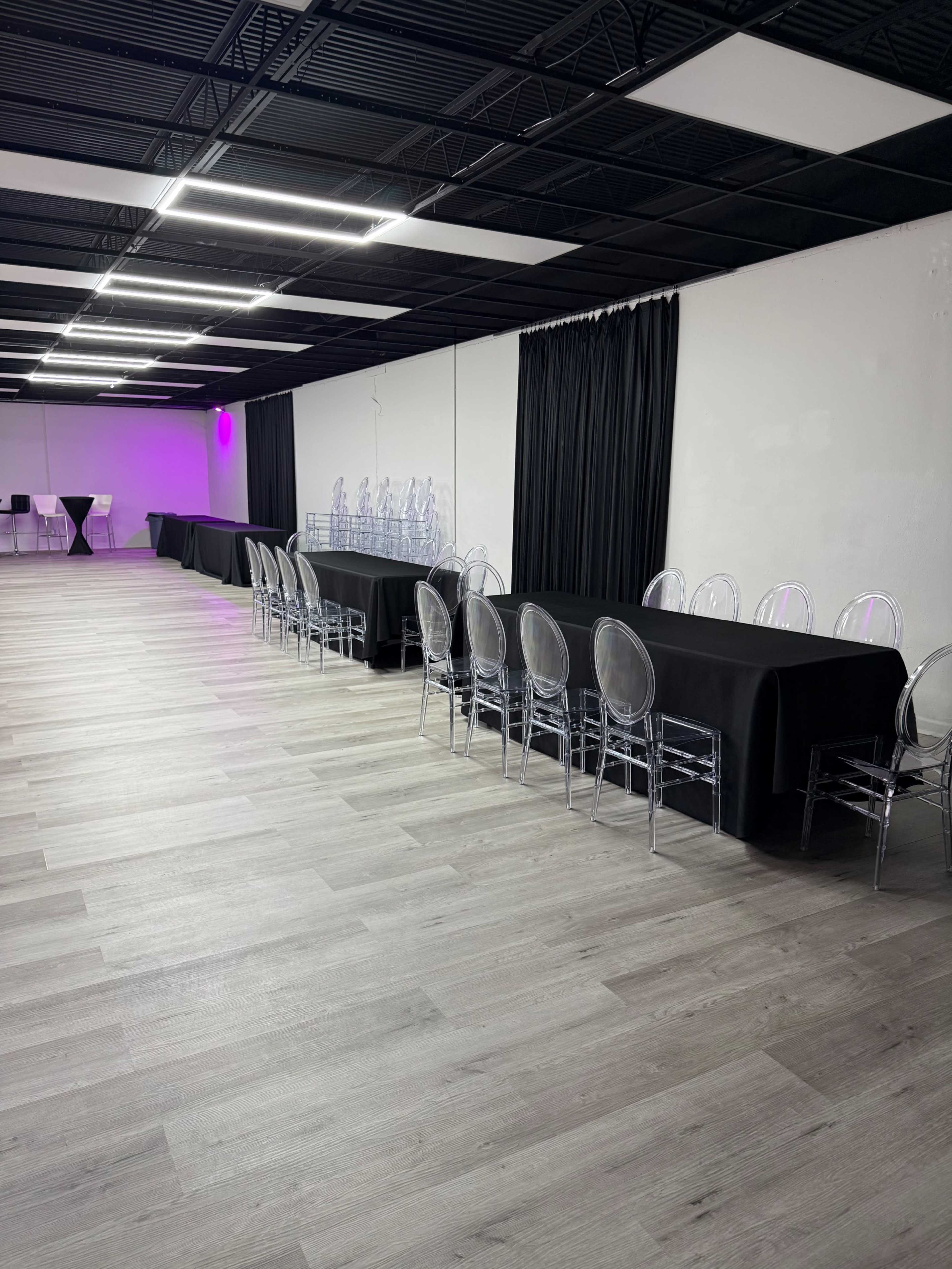 The image shows a spacious indoor venue with long tables covered in black cloth and clear plastic chairs arranged along one side, while purple lighting illuminates part of the room.