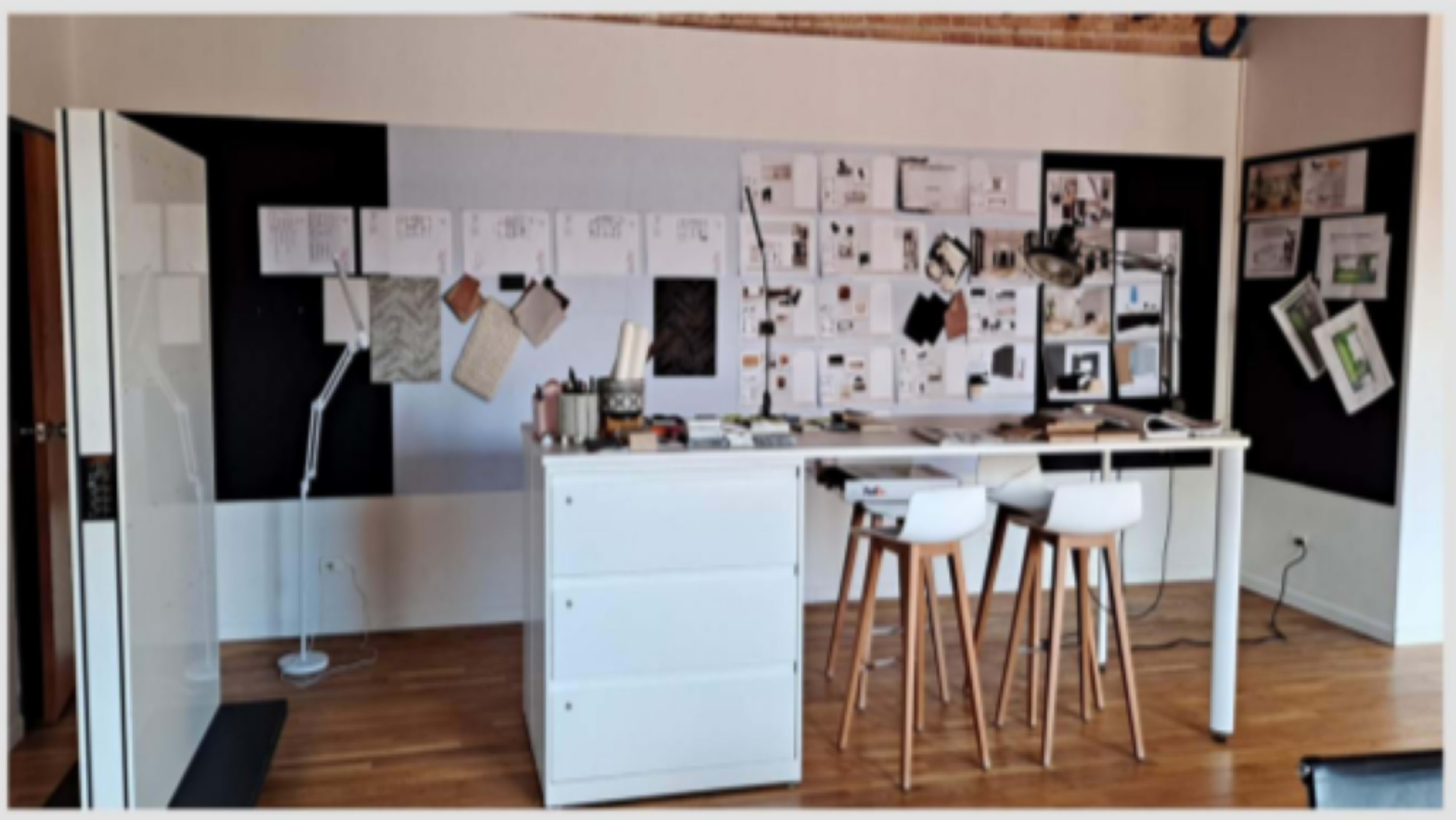 A minimalist workspace with a central table, white stools, and a wall covered in various papers and design materials.