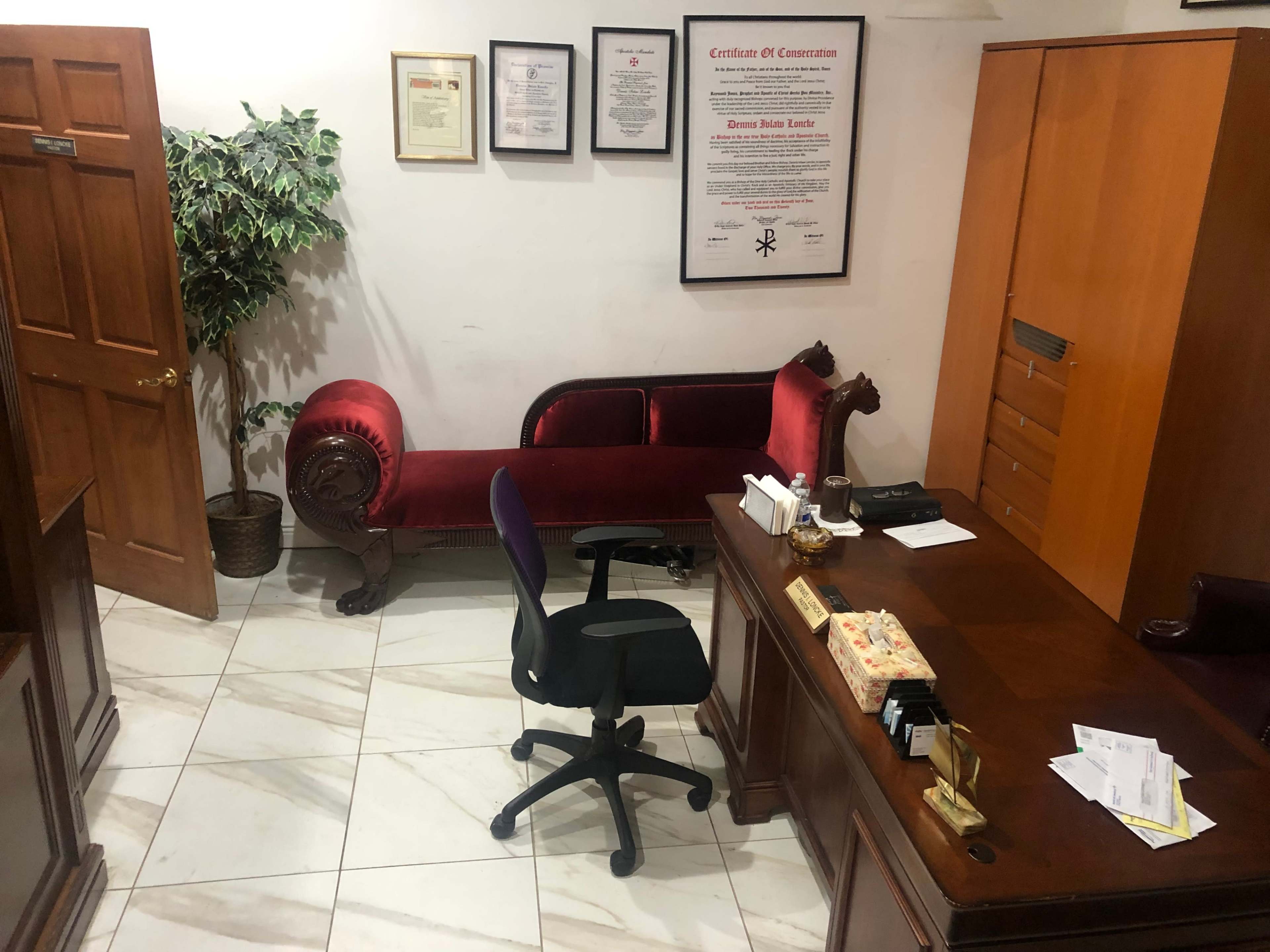 An office with a wooden desk, an office chair, a red upholstered sofa, and a decorative plant in the corner.
