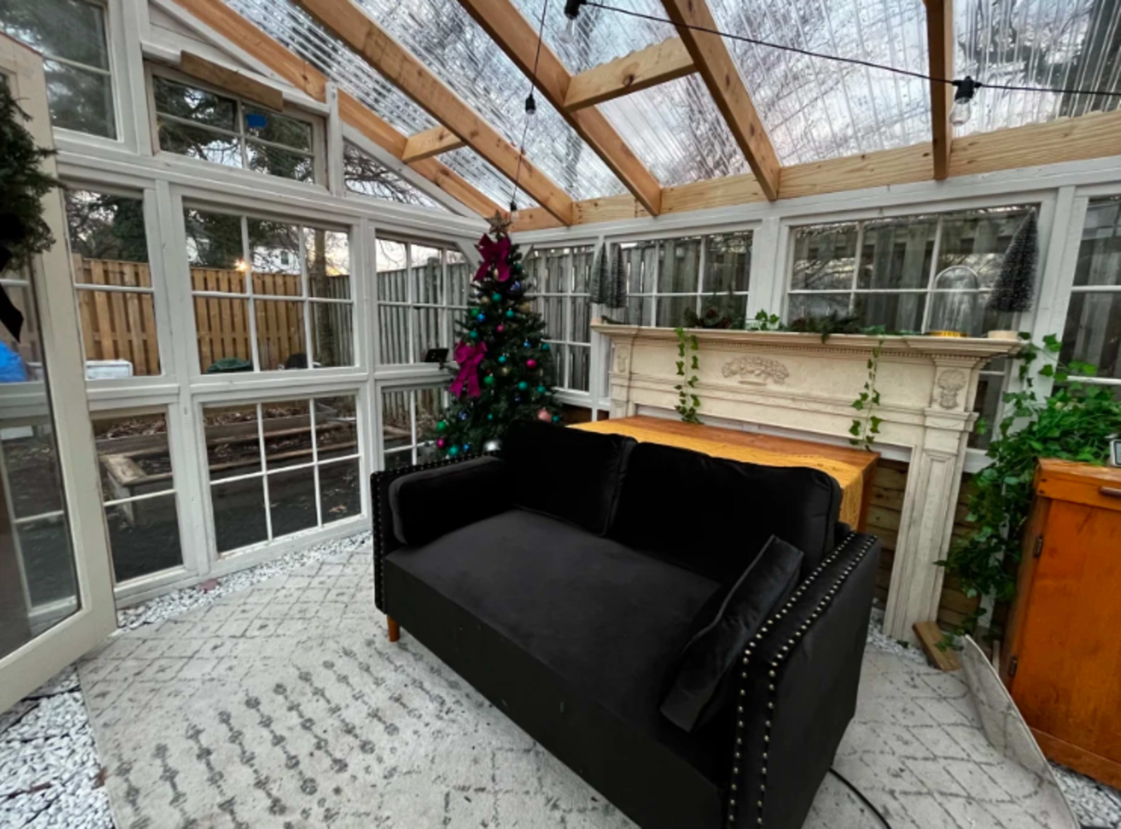 A cozy sunroom featuring a black couch, a Christmas tree, a decorative fireplace, and large windows with a wooden ceiling.