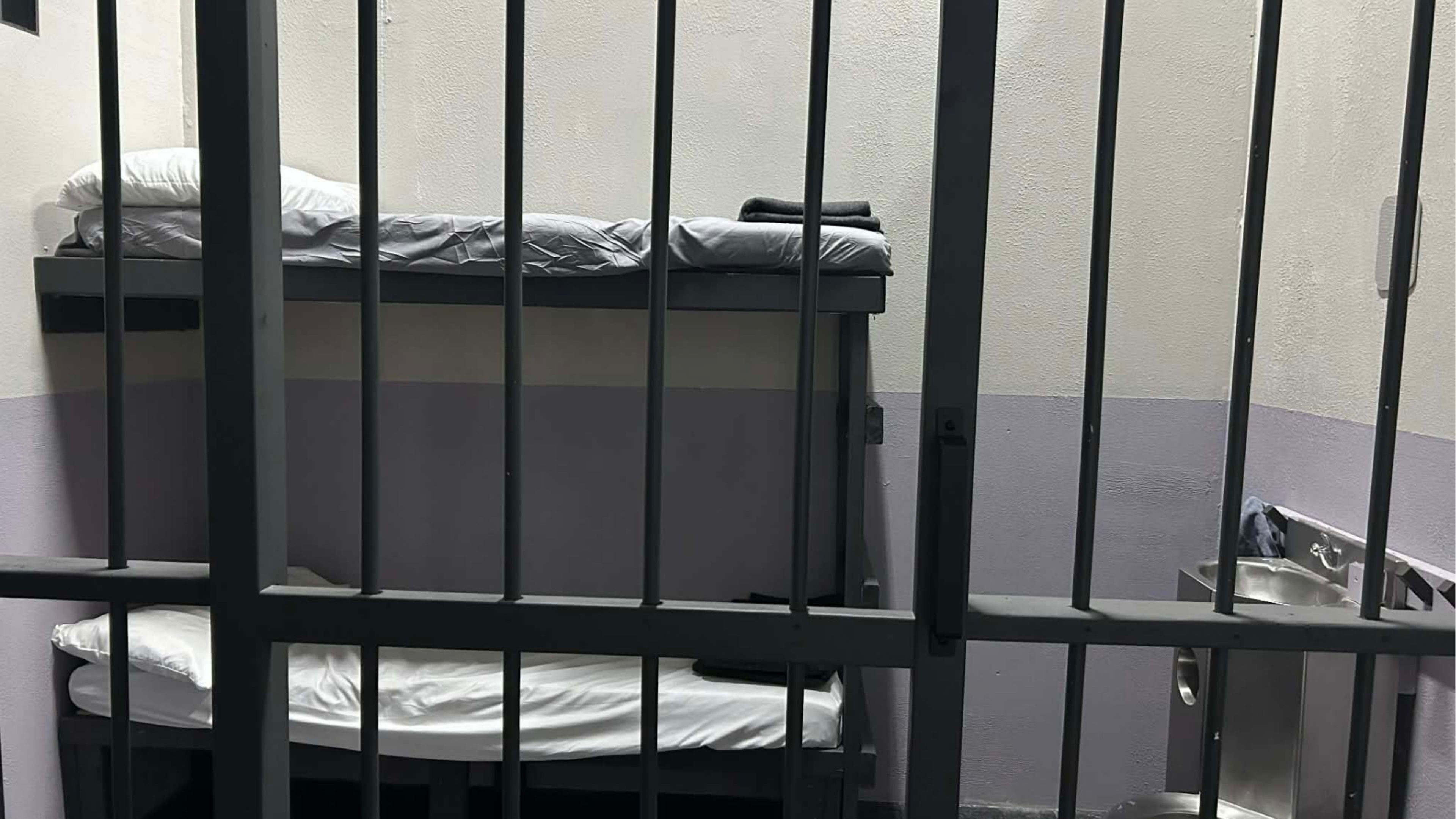The image shows the interior of a small jail cell with two metal-framed beds and a wall-mounted sink.