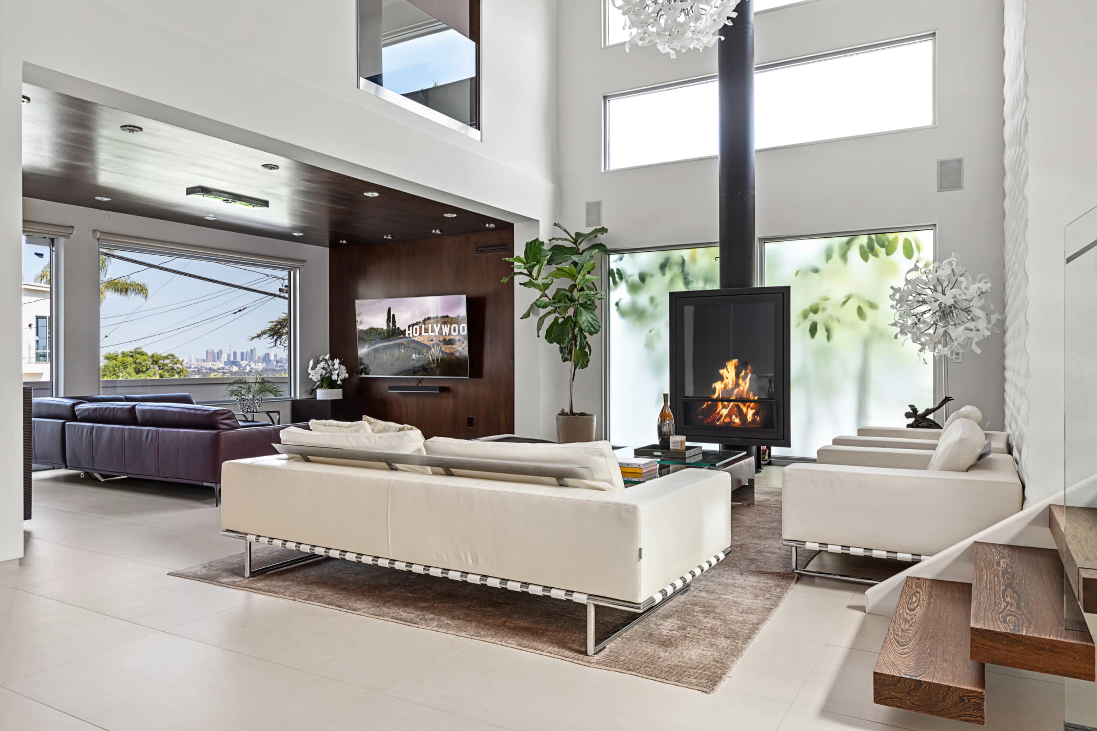 The image shows a modern living room with two white sofas, a dark wood accent wall featuring a television, a fireplace, and large windows that provide a view of the city.