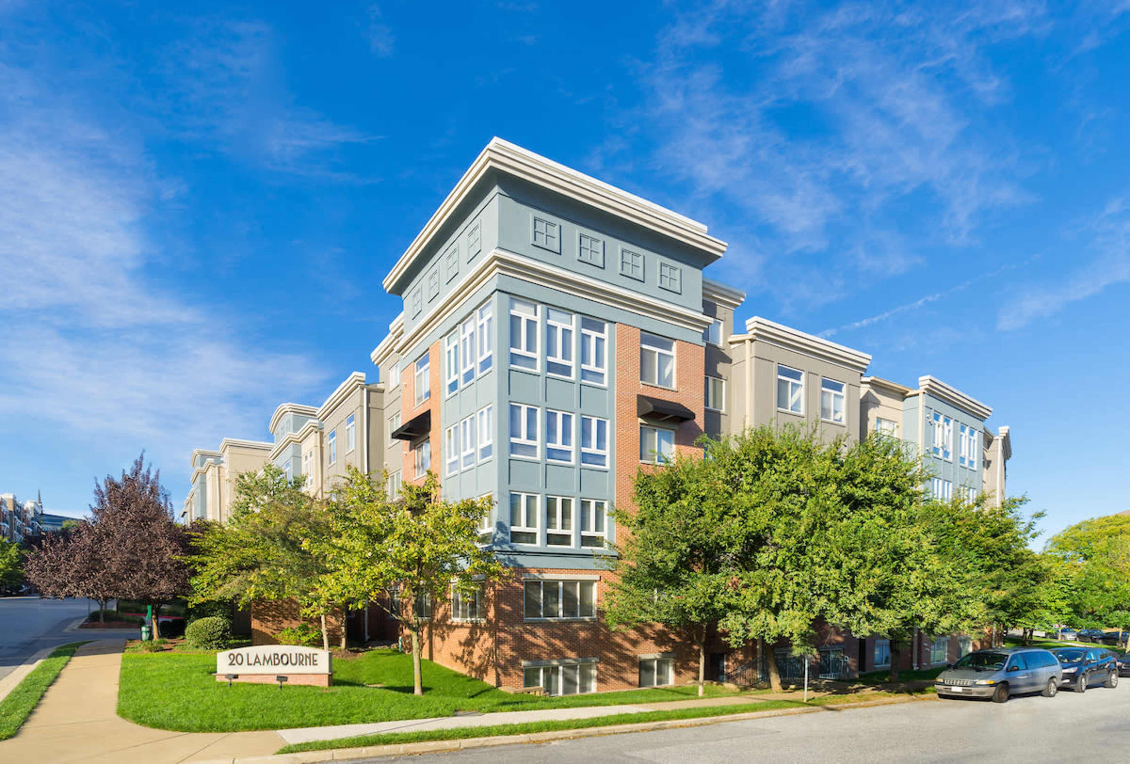 The image shows a modern, multi-story apartment building with a sign reading "20 Landbourne" in front, surrounded by trees and a clear blue sky.