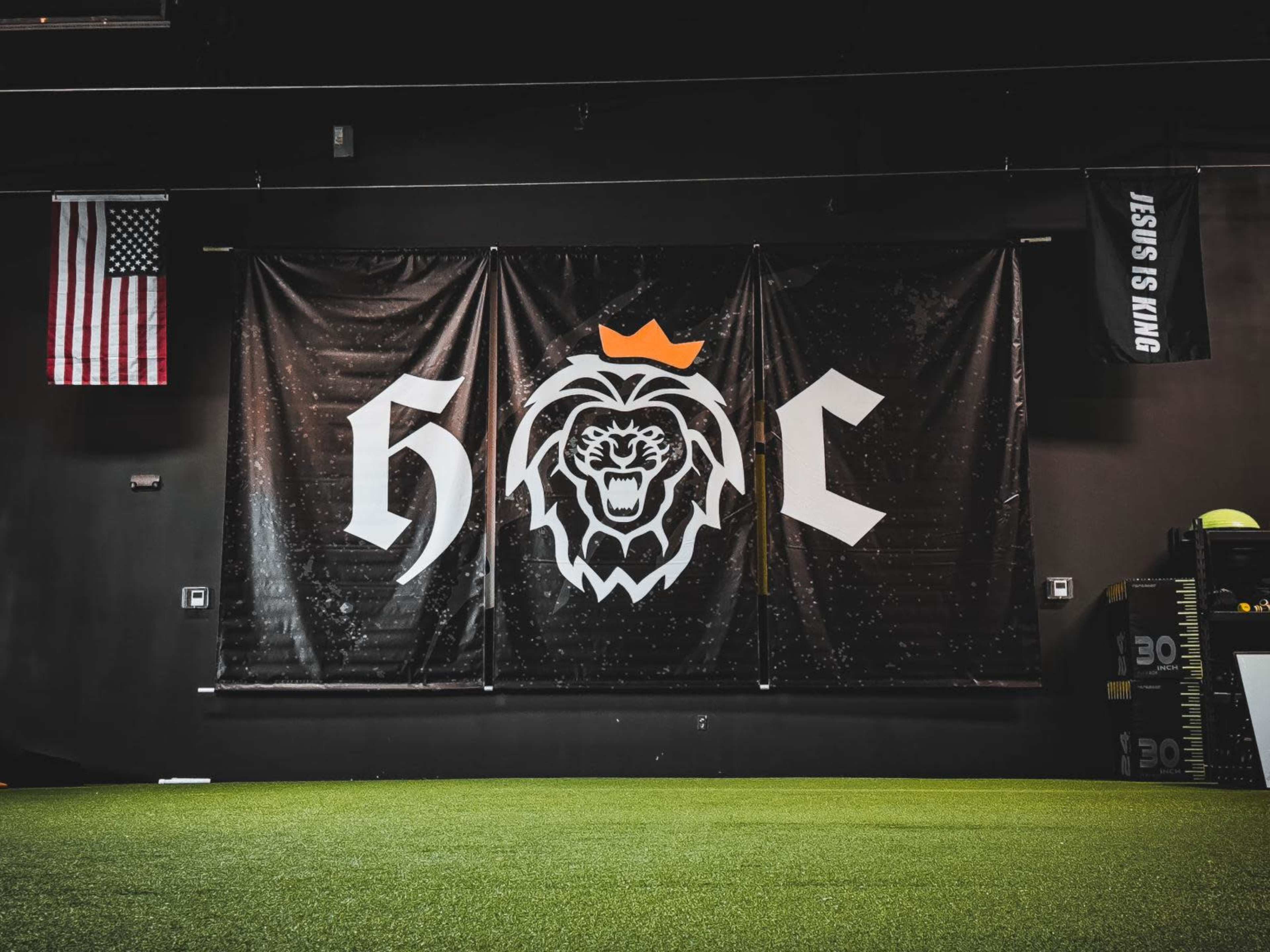 A large black banner with a lion's head logo and the letters "H" and "C" in a gym setting, flanked by an American flag on the left and a banner reading "JESUS IS KING" on the right.