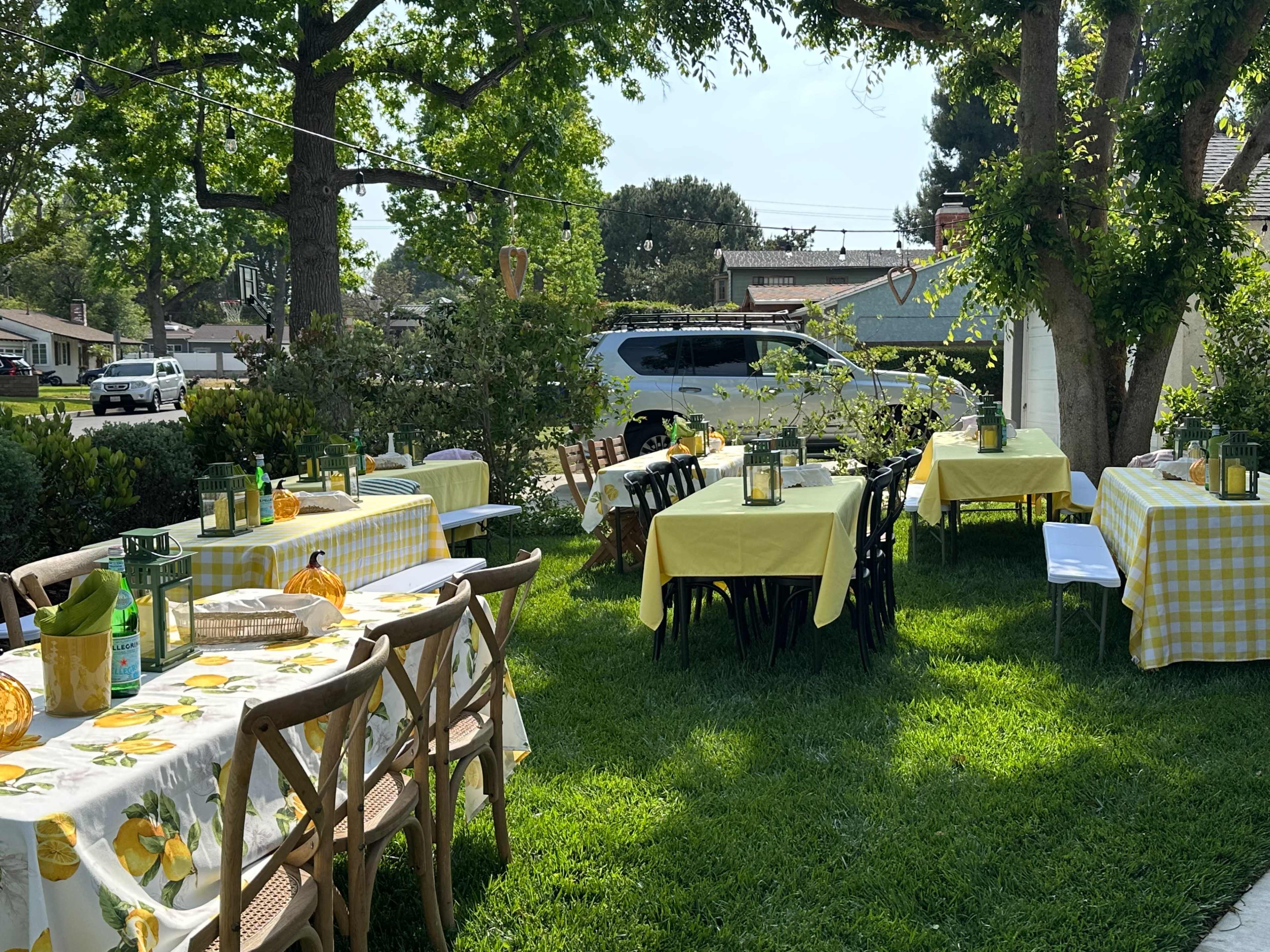 The image shows a garden setting with several tables covered in yellow tablecloths, surrounded by greenery and enhancing the atmosphere of an outdoor gathering.
