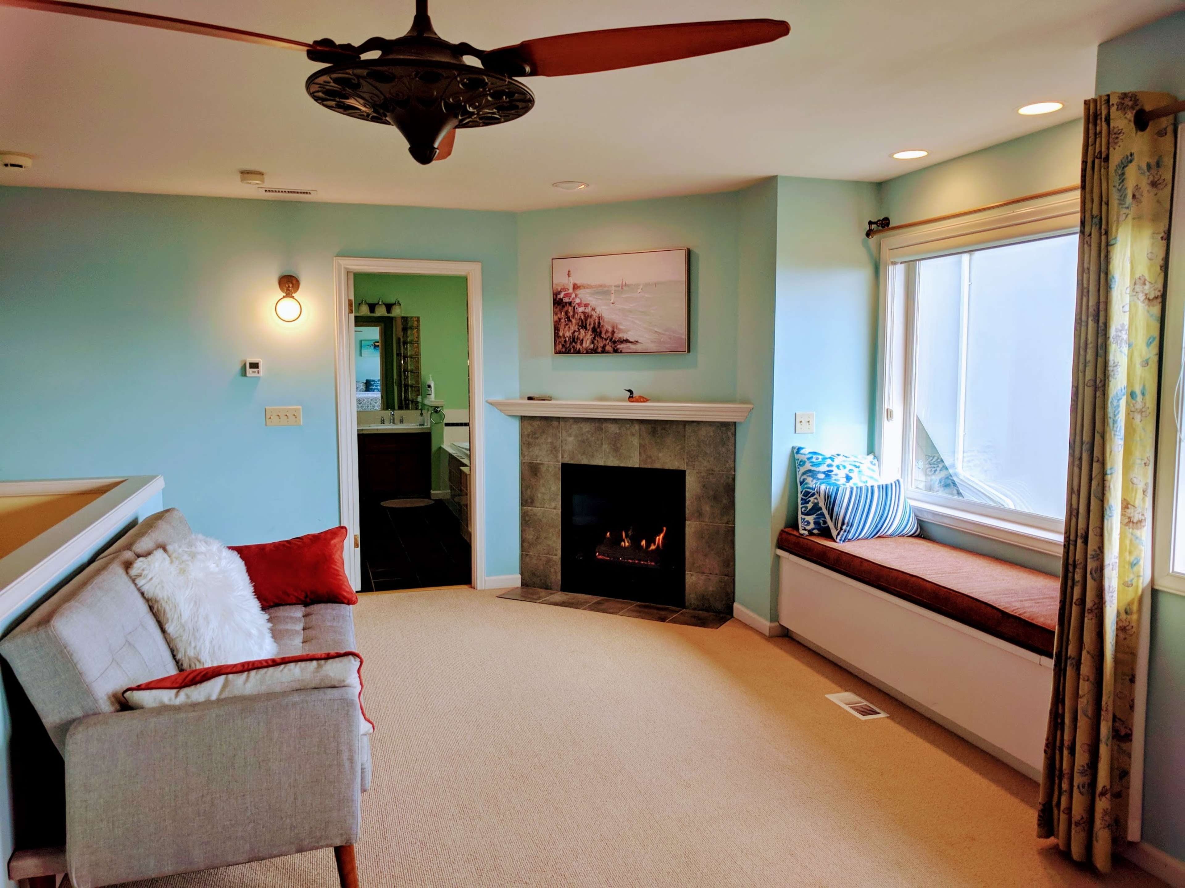 A cozy living room features a fireplace, a window seat, and a gray sofa, with light blue walls and natural light entering from the window.
