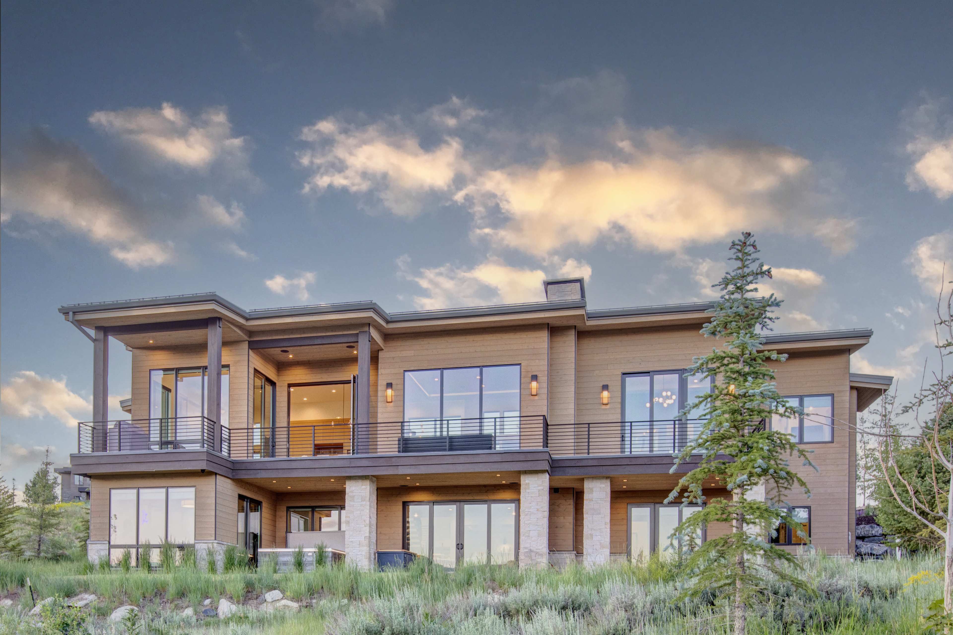 A modern two-story house with large windows, a balcony, and surrounding greenery against a cloudy sky.