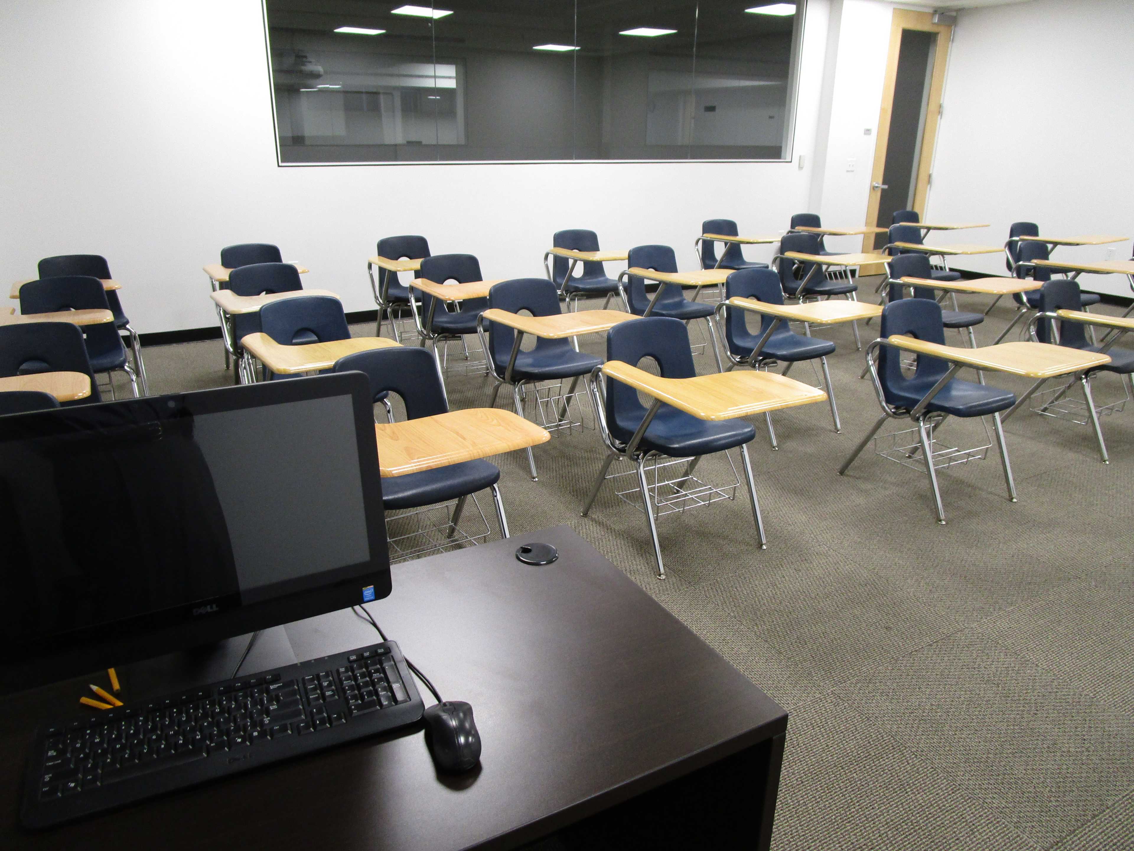 A classroom is arranged with rows of empty desks and a computer on a desk at the front.