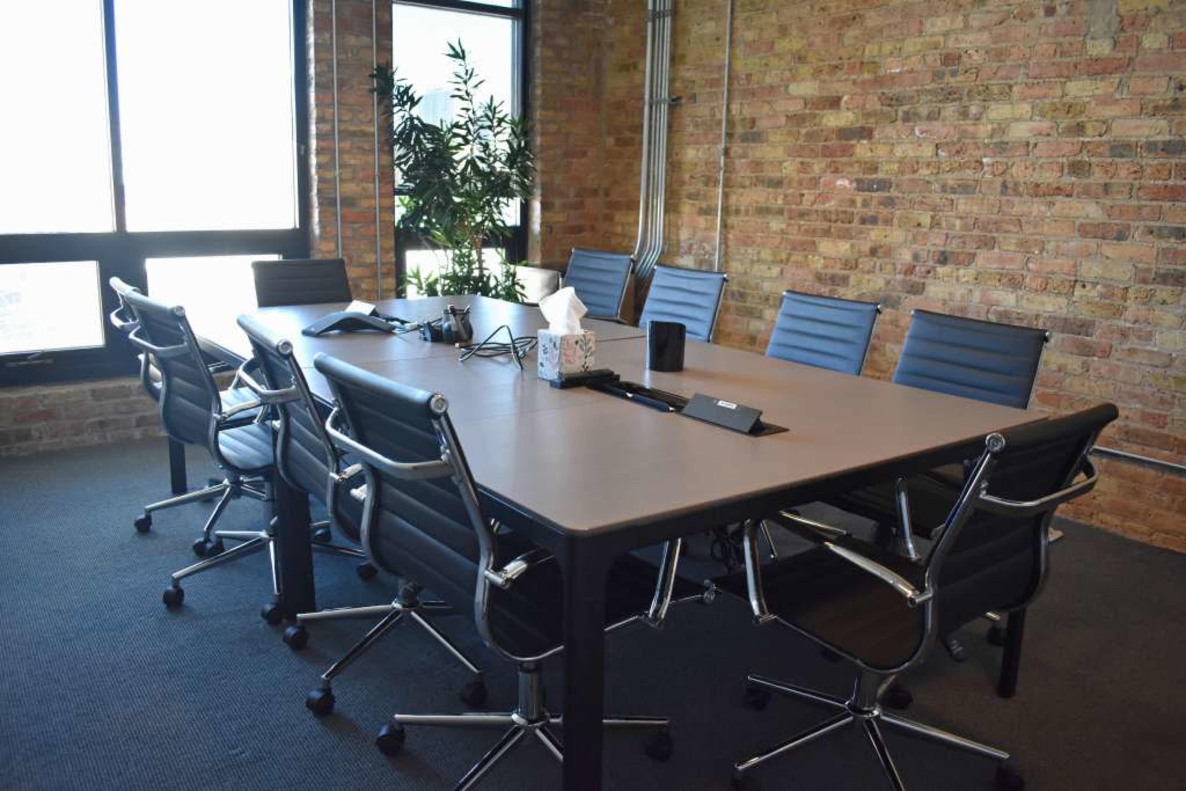 A modern conference room features a large rectangular table surrounded by ergonomic chairs, with windows and exposed brick walls.