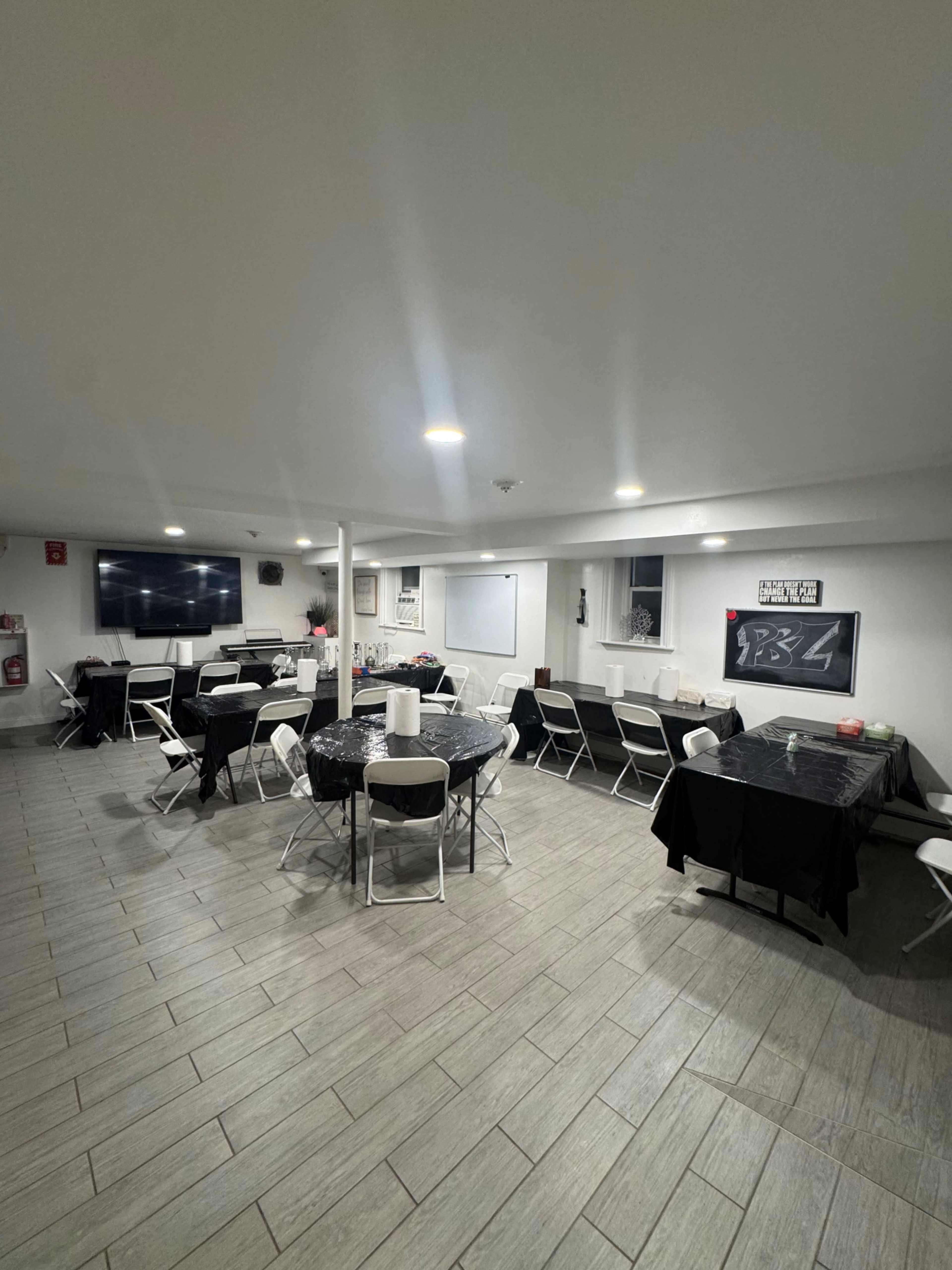 The image shows a spacious room set up for an event, featuring several round tables covered with black tablecloths and surrounding chairs, along with a large TV and whiteboard at one end.