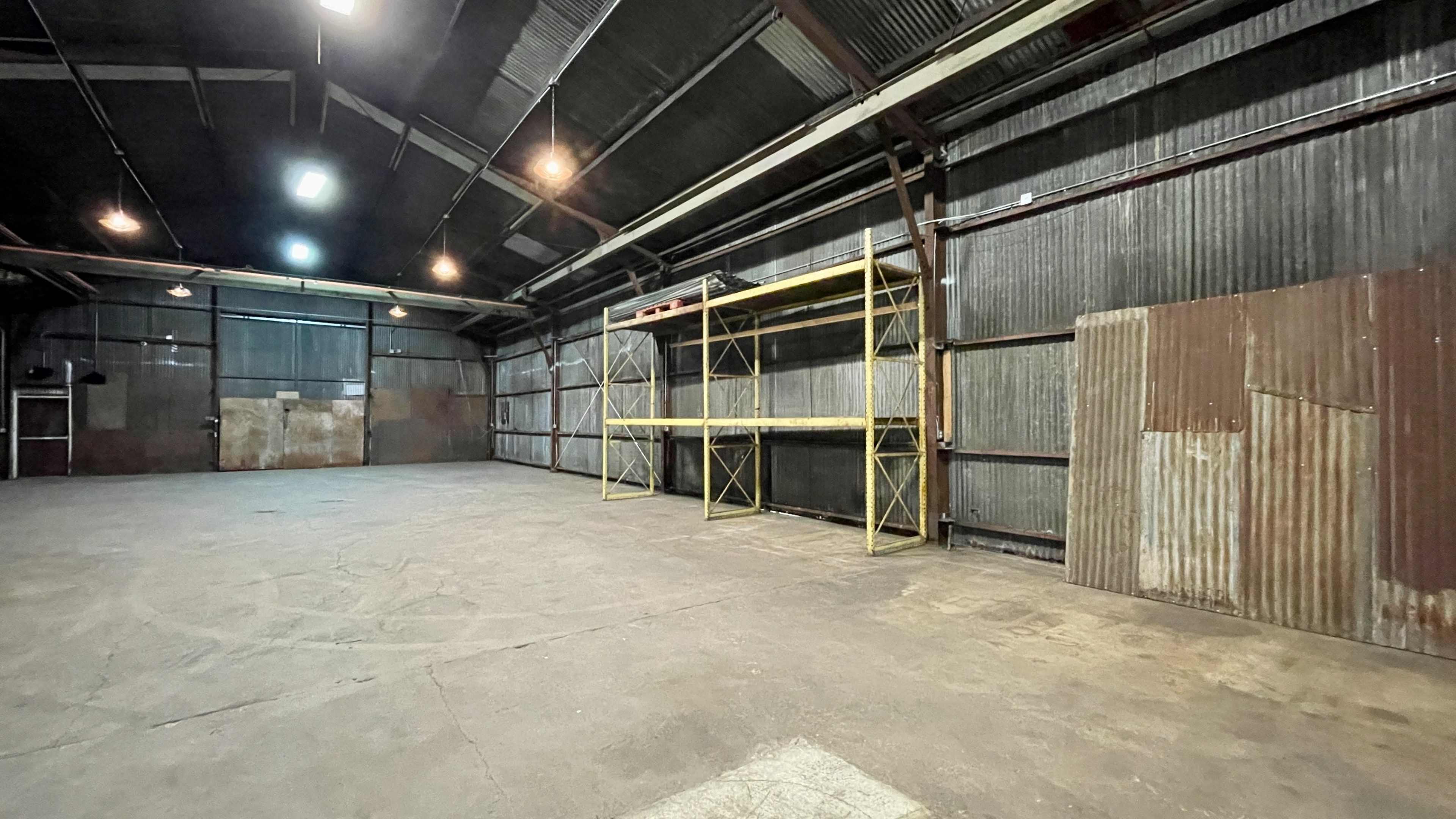 A spacious, industrial warehouse with a concrete floor, corrugated metal walls, and a yellow shelving unit along one side.