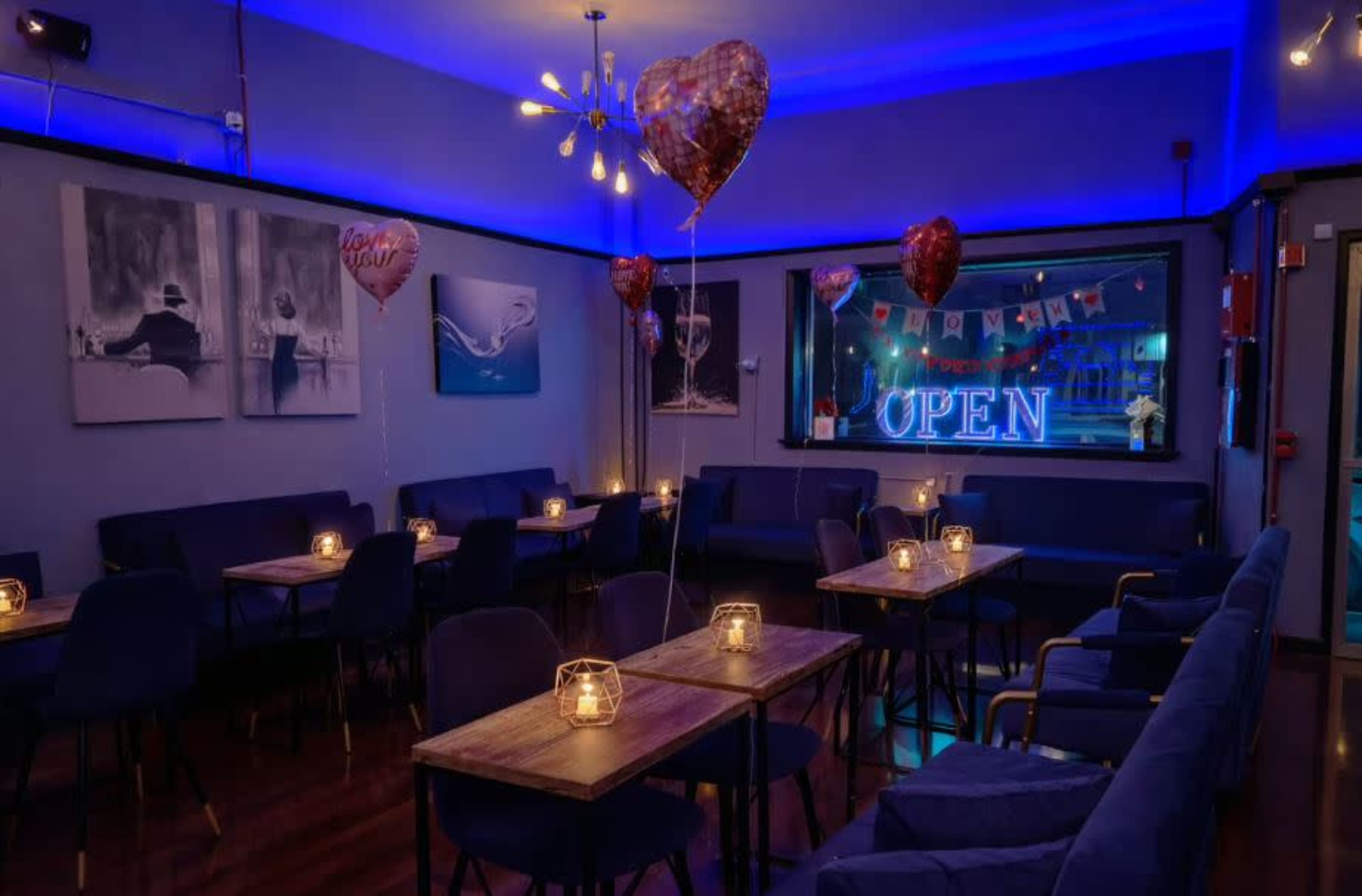 The image shows a cozy café setting with illuminated tables, heart-shaped balloons, and a blue-toned atmosphere.