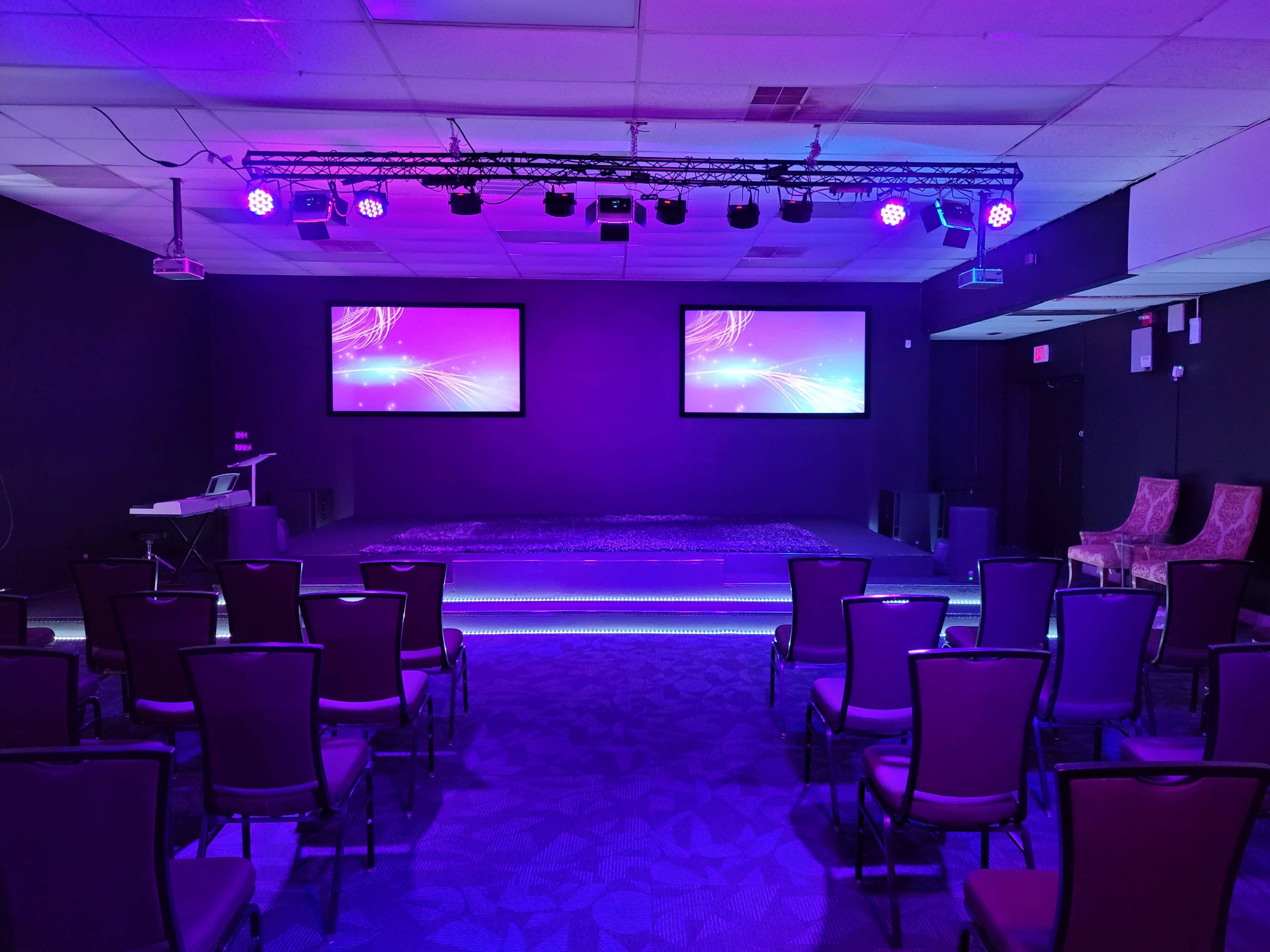 A stage with two screens is set in a dimly lit room, surrounded by rows of chairs and illuminated by purple lights.