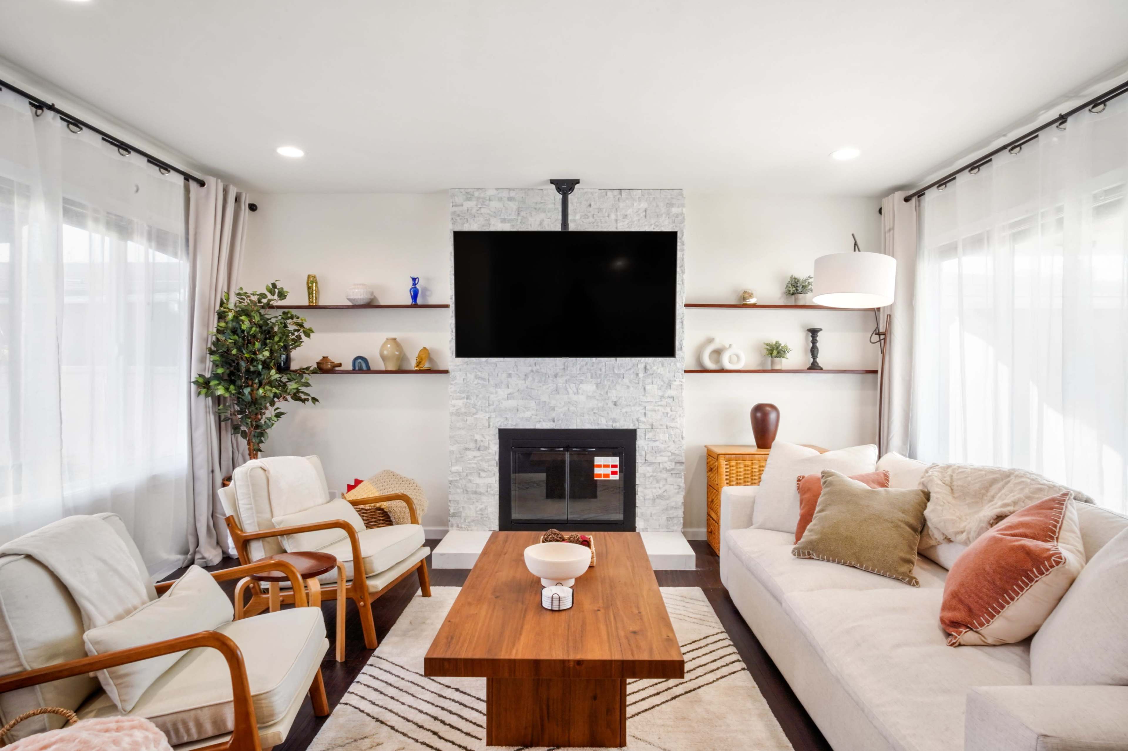 The living room features a white sofa, wooden coffee table, and a wall-mounted TV above a stone fireplace, with shelves displaying decorative items.
