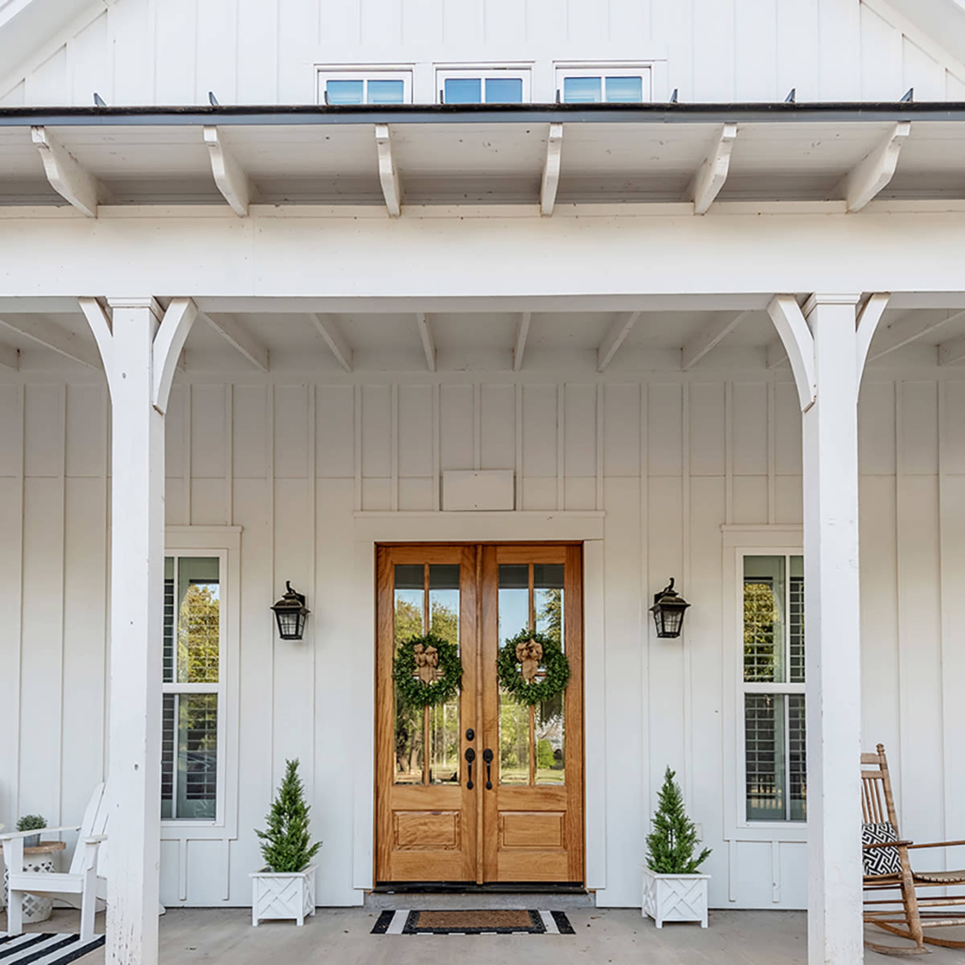 A white farmhouse-style entrance features double wooden doors adorned with wreaths, flanked by small potted evergreen plants and set under a covered porch.