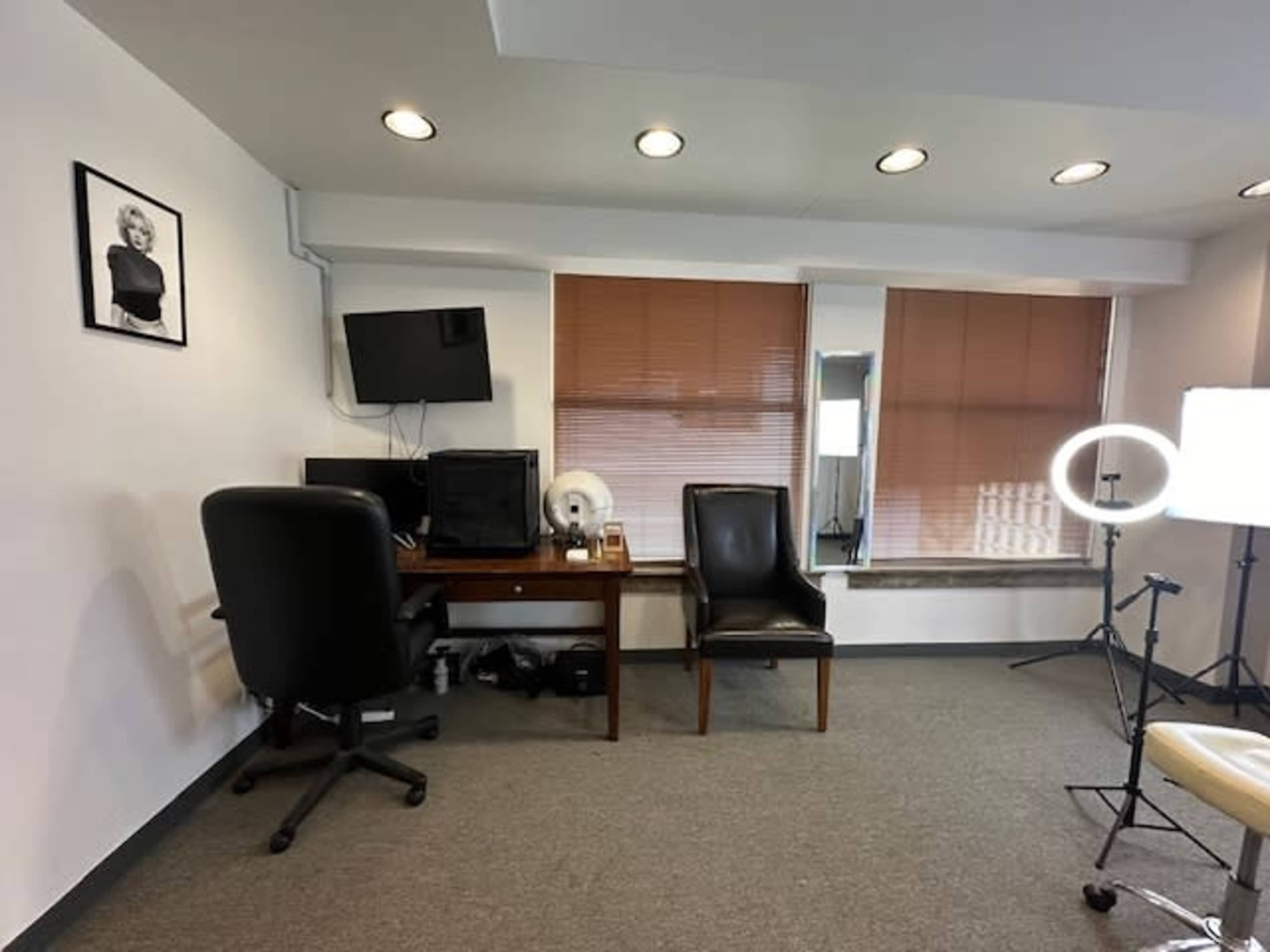 The image shows a simple office space with a desk, two chairs, a computer monitor, and large windows featuring blinds.