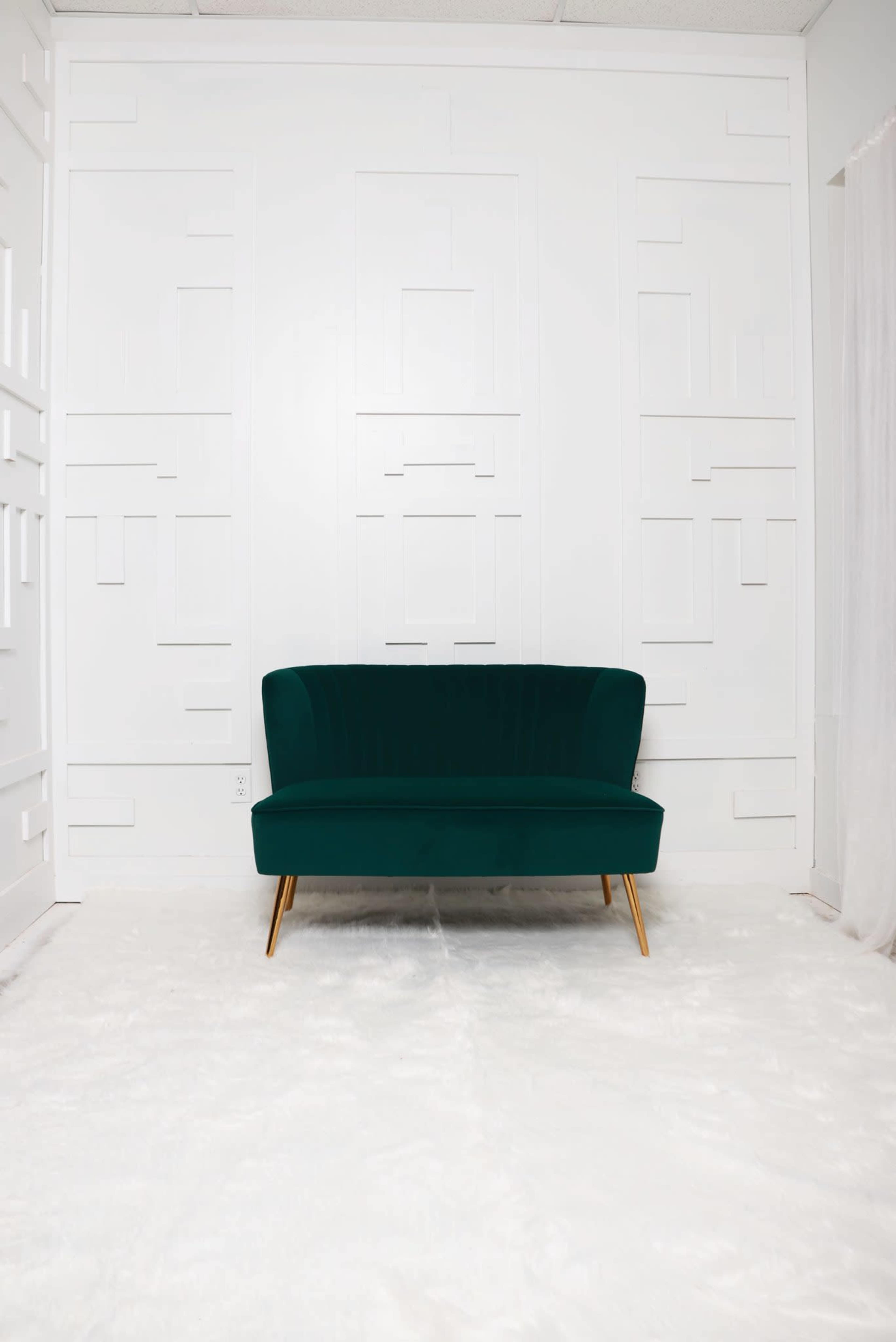 A green sofa with gold legs is positioned against a white textured wall in an empty room with a light-colored floor.