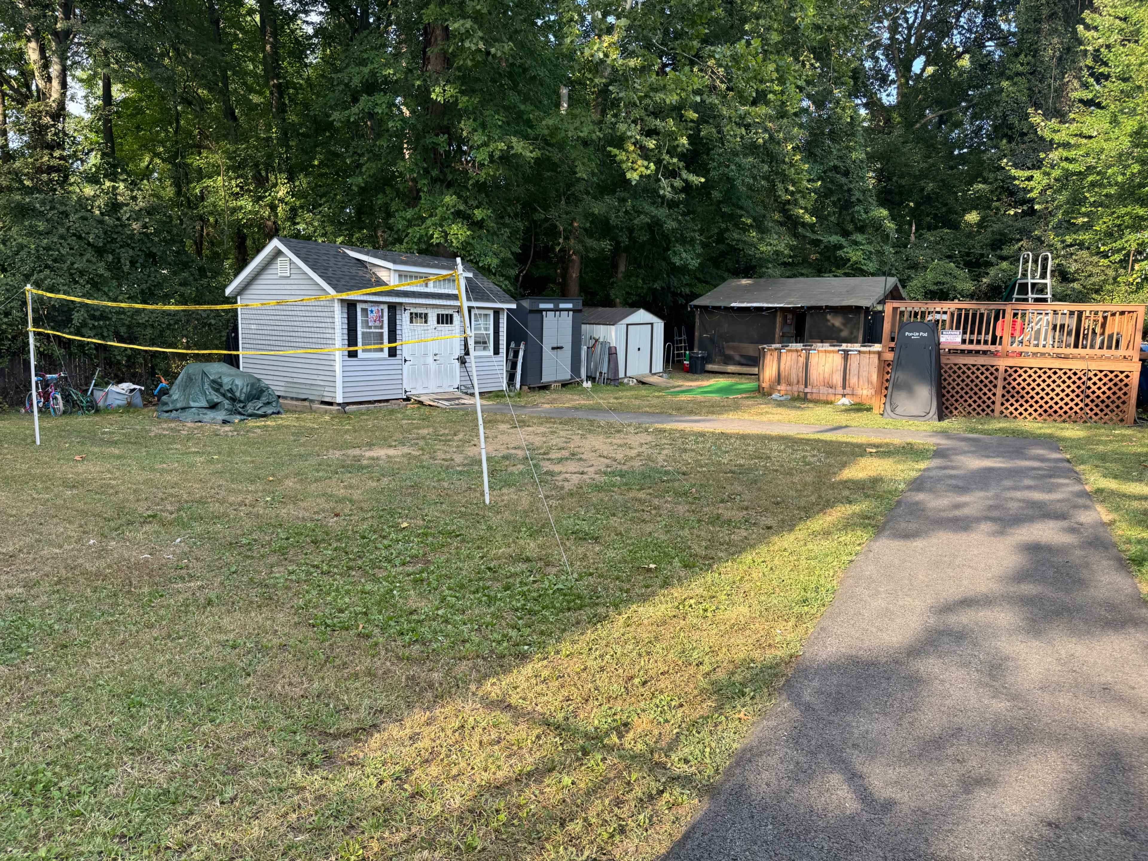 The image shows a backyard with a small white shed, a gray storage unit, a covered area, a wooden deck with a pool, and a volleyball net set up in the grassy space.