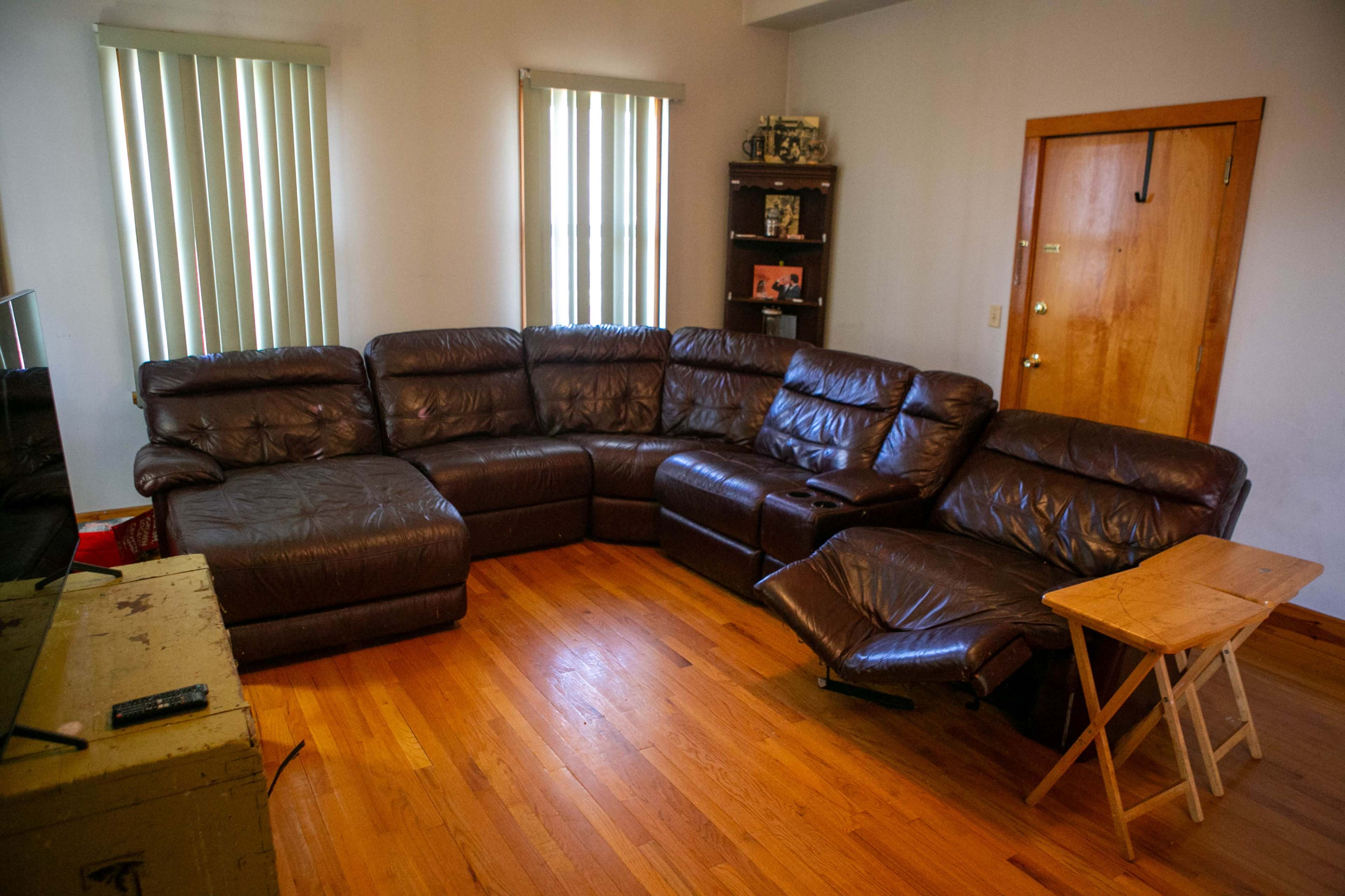 The image shows a spacious living room with a brown sectional sofa, a small wooden side table, and a wooden bookshelf near the door.