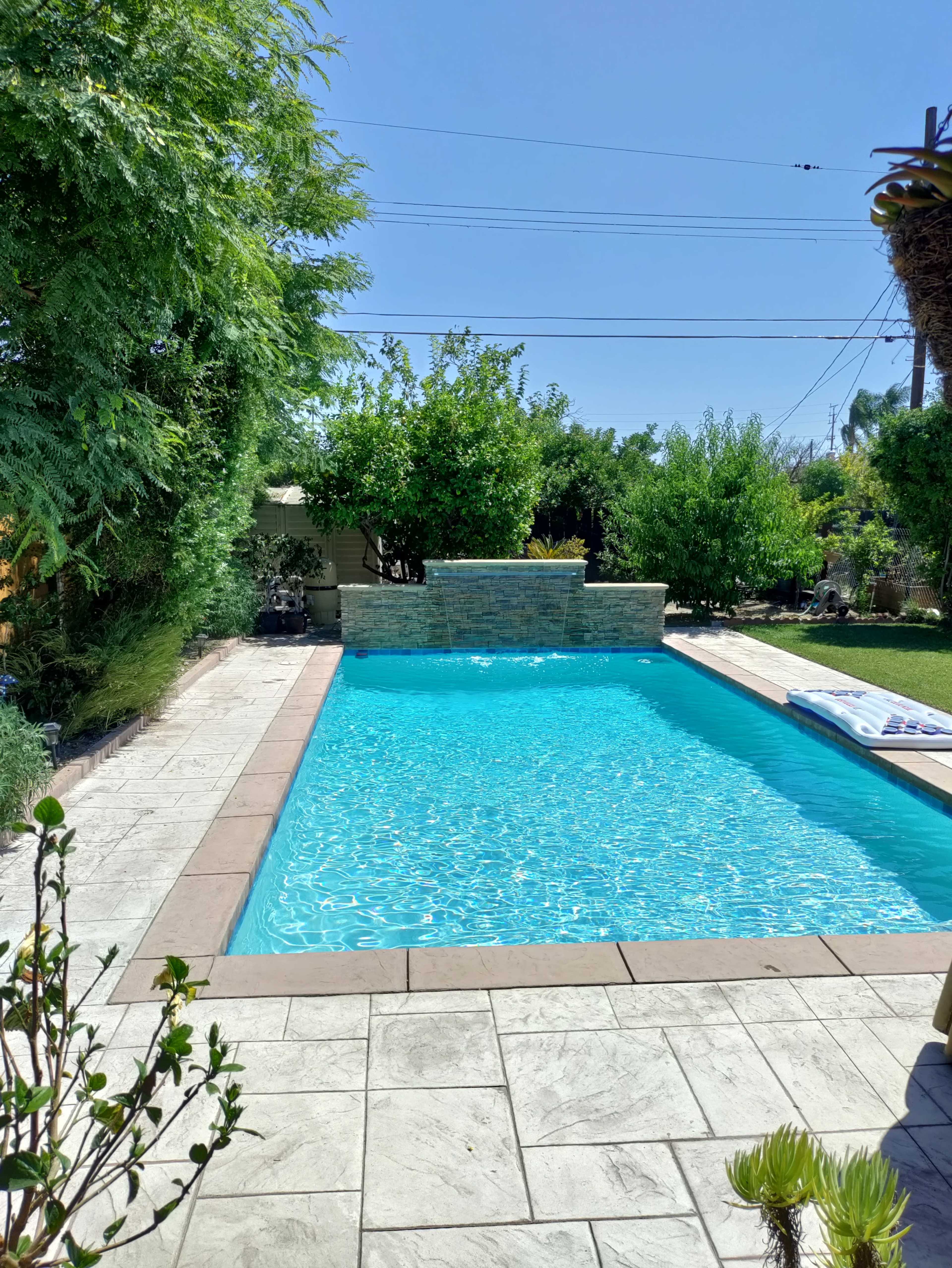 The image shows a rectangular swimming pool surrounded by a stone patio and landscaped greenery under a clear blue sky.