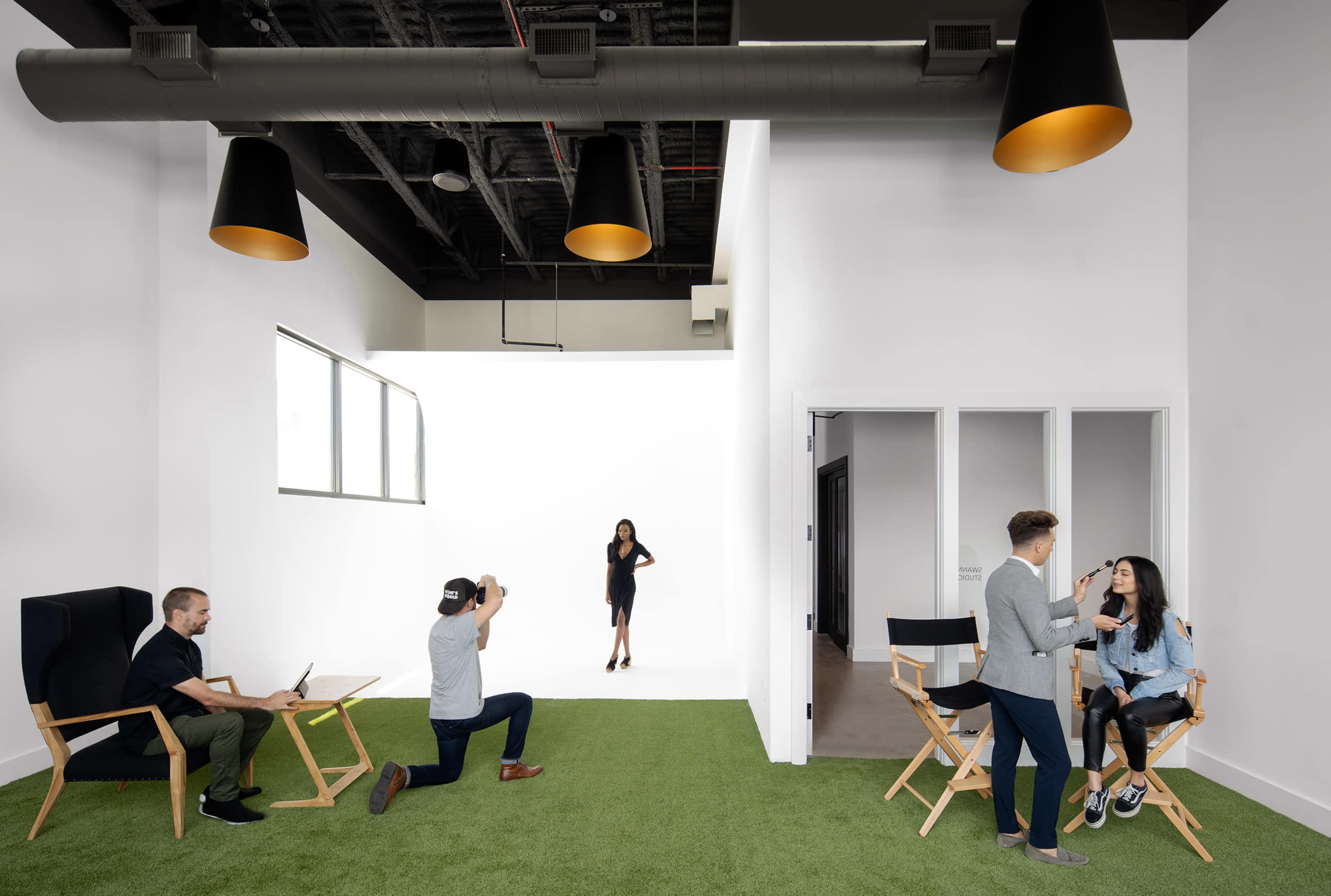 A modern studio space features a model posing against a white backdrop while a photographer captures her image, and two groups are engaged in conversation nearby.