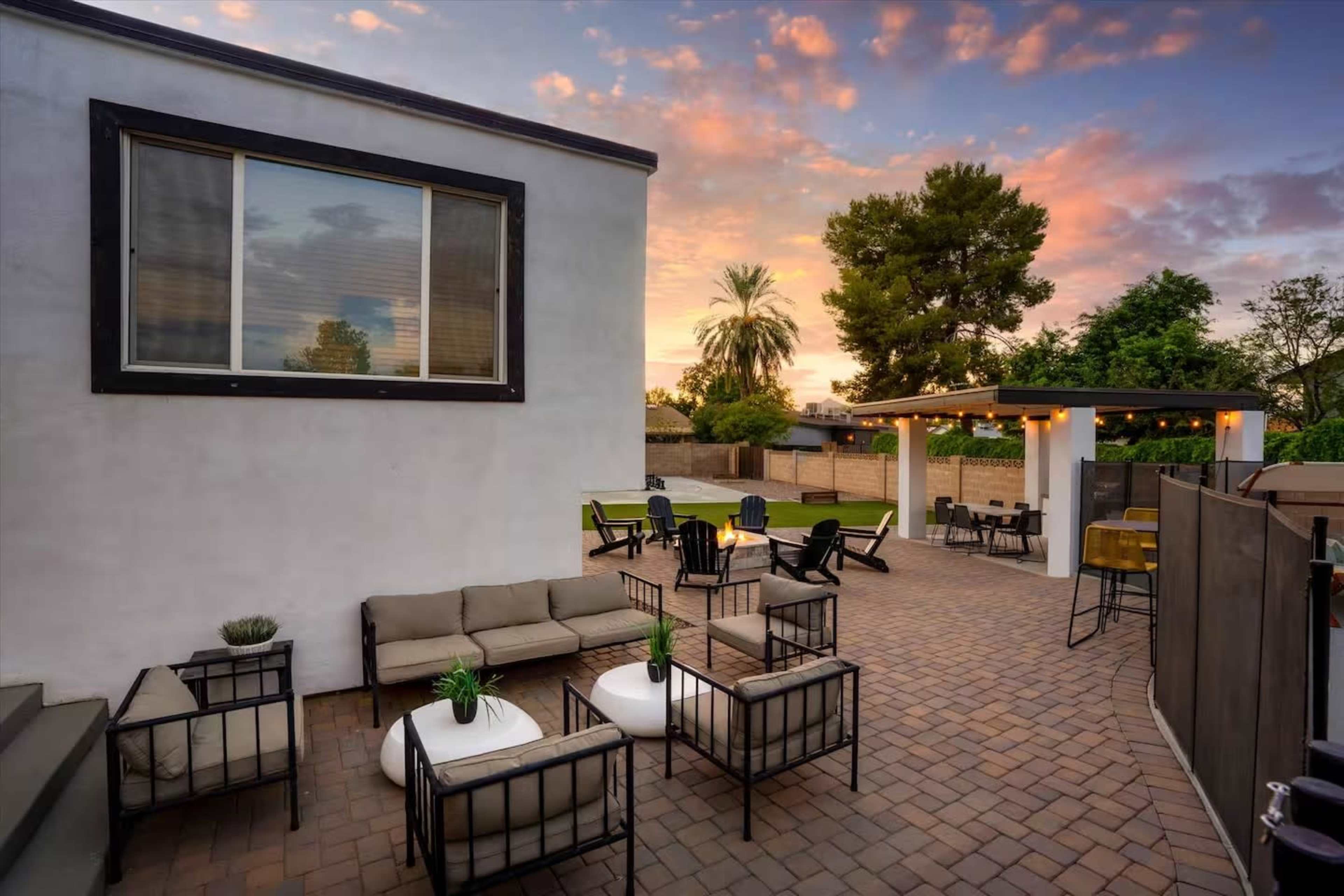 The image shows a landscaped outdoor patio area with seating arrangements, tables, and a backdrop of a sunset sky.