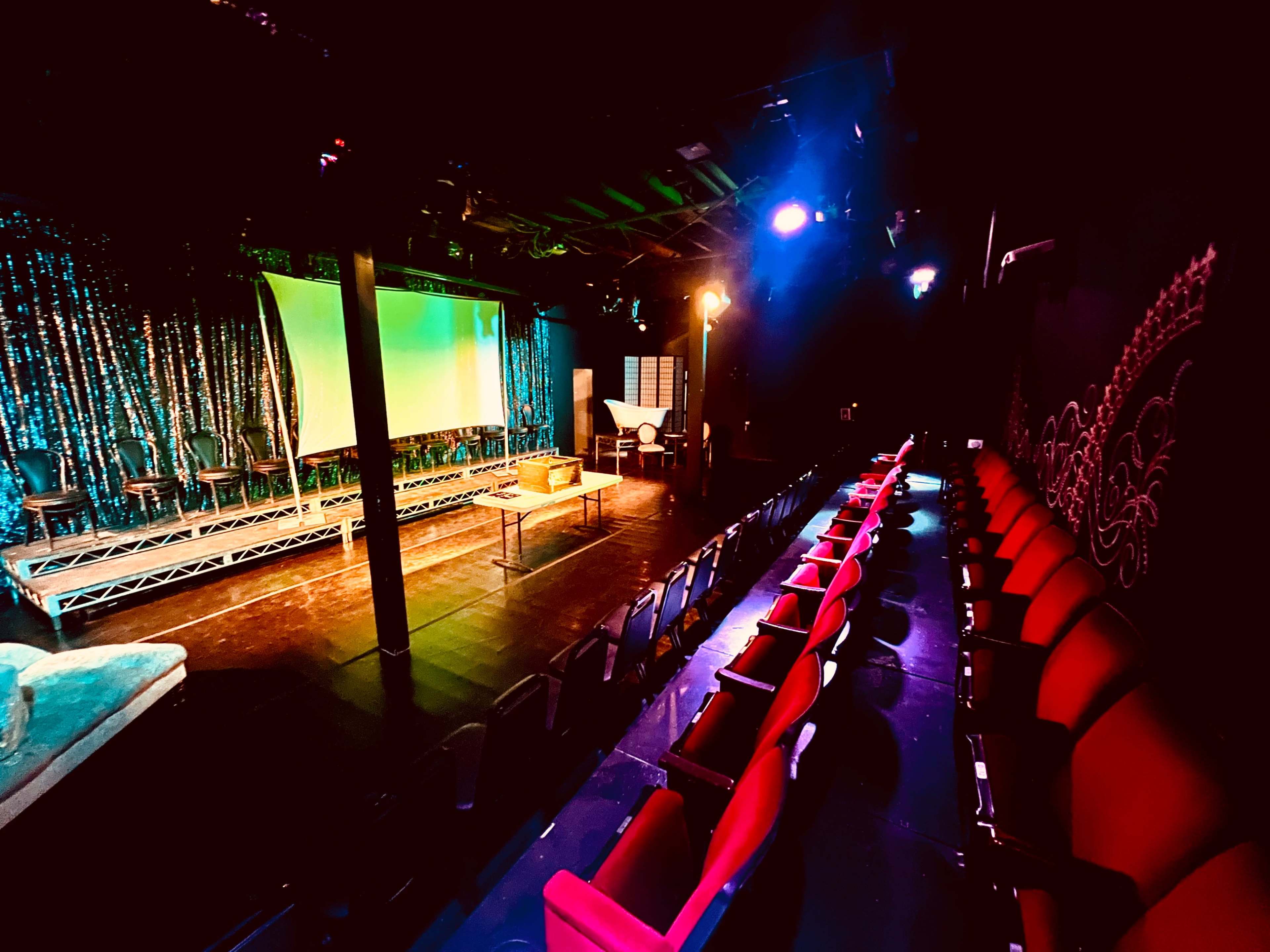 A dimly lit theater with red seating facing a stage that has a large screen and a backdrop adorned with shimmering curtains.