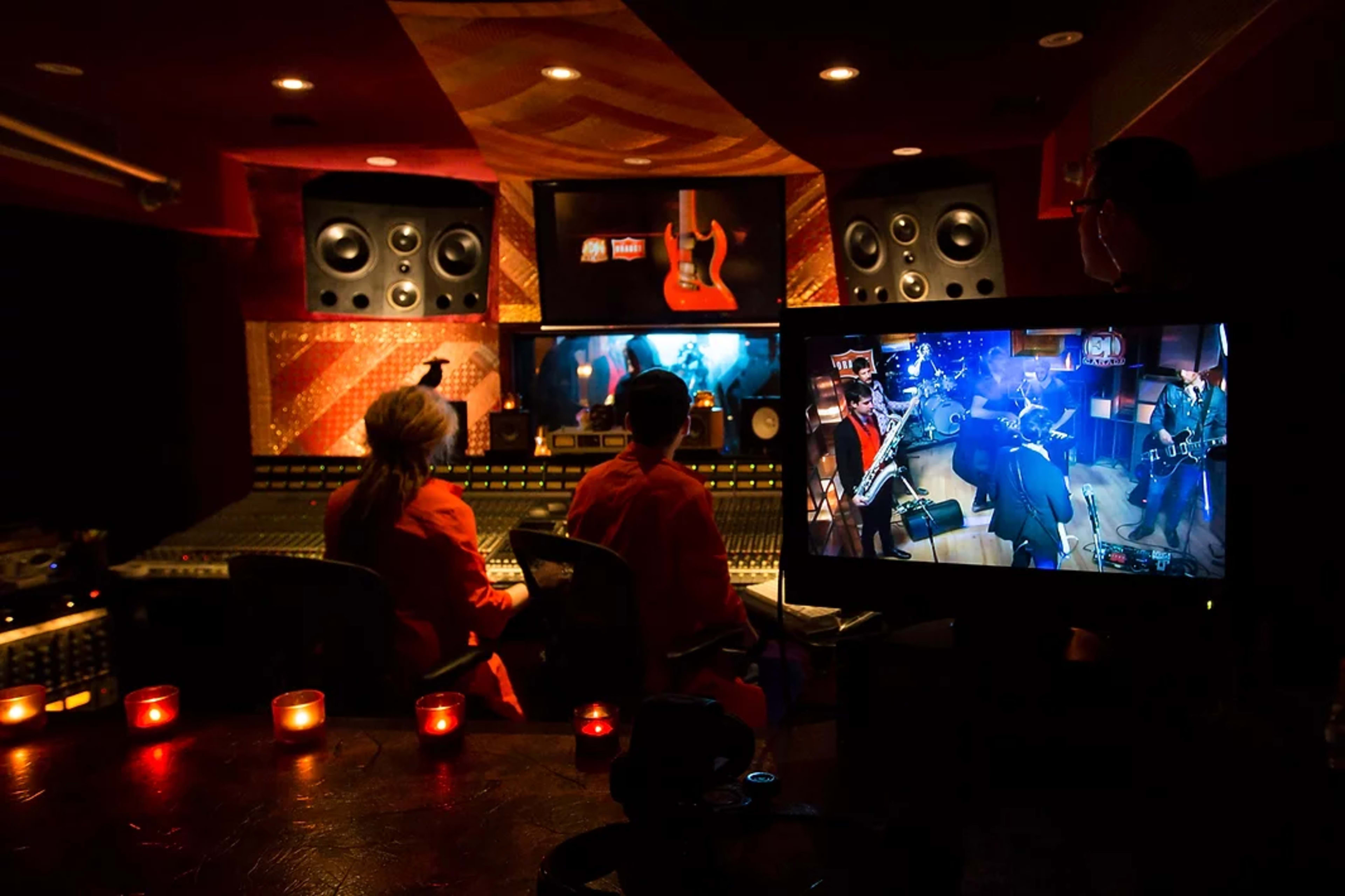 Two people sit in a sound studio, surrounded by mixing equipment, while a live band plays on a screen in front of them.