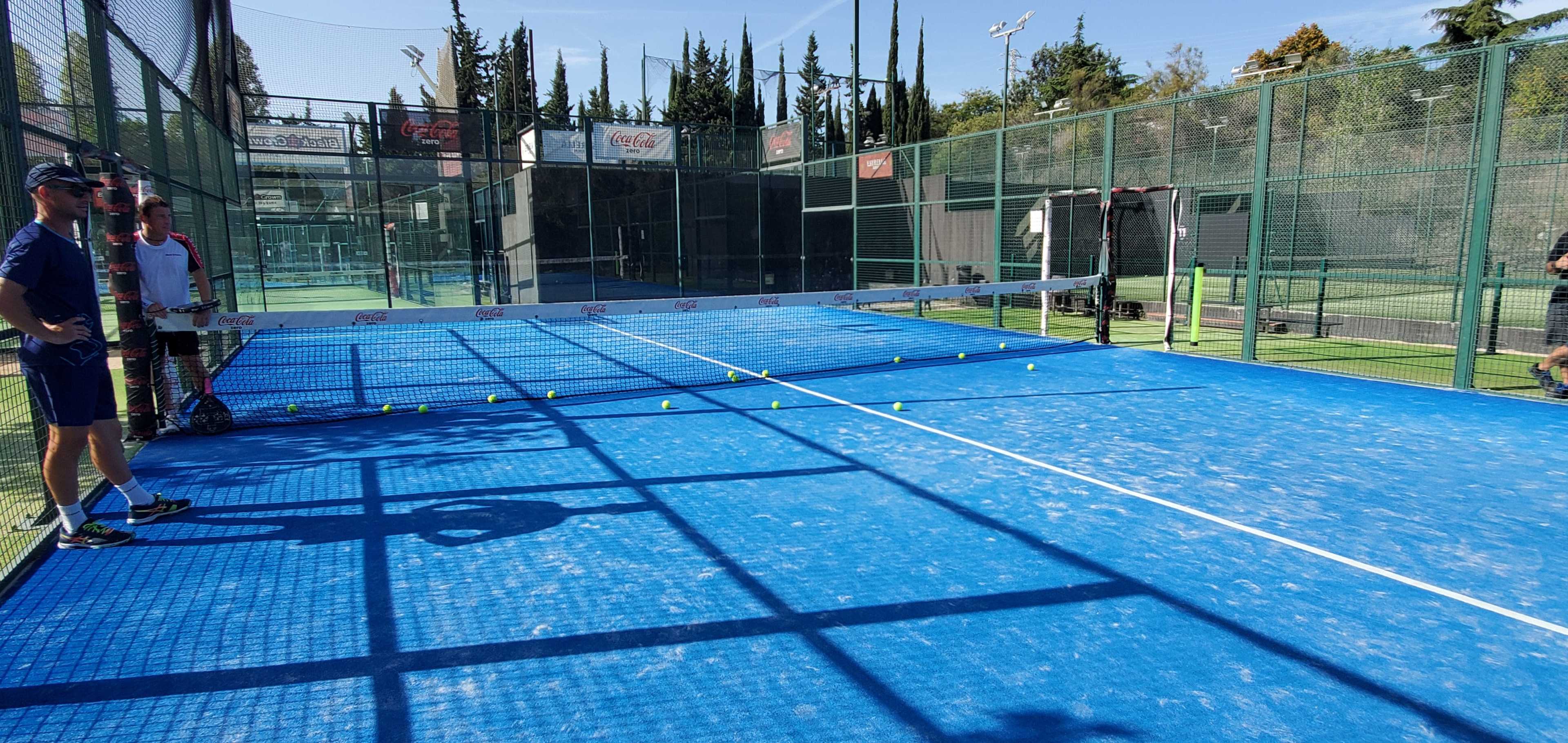 A blue padel court with several balls arranged along the center line, and two players, one in a blue uniform and another in a white shirt, standing beside the court.