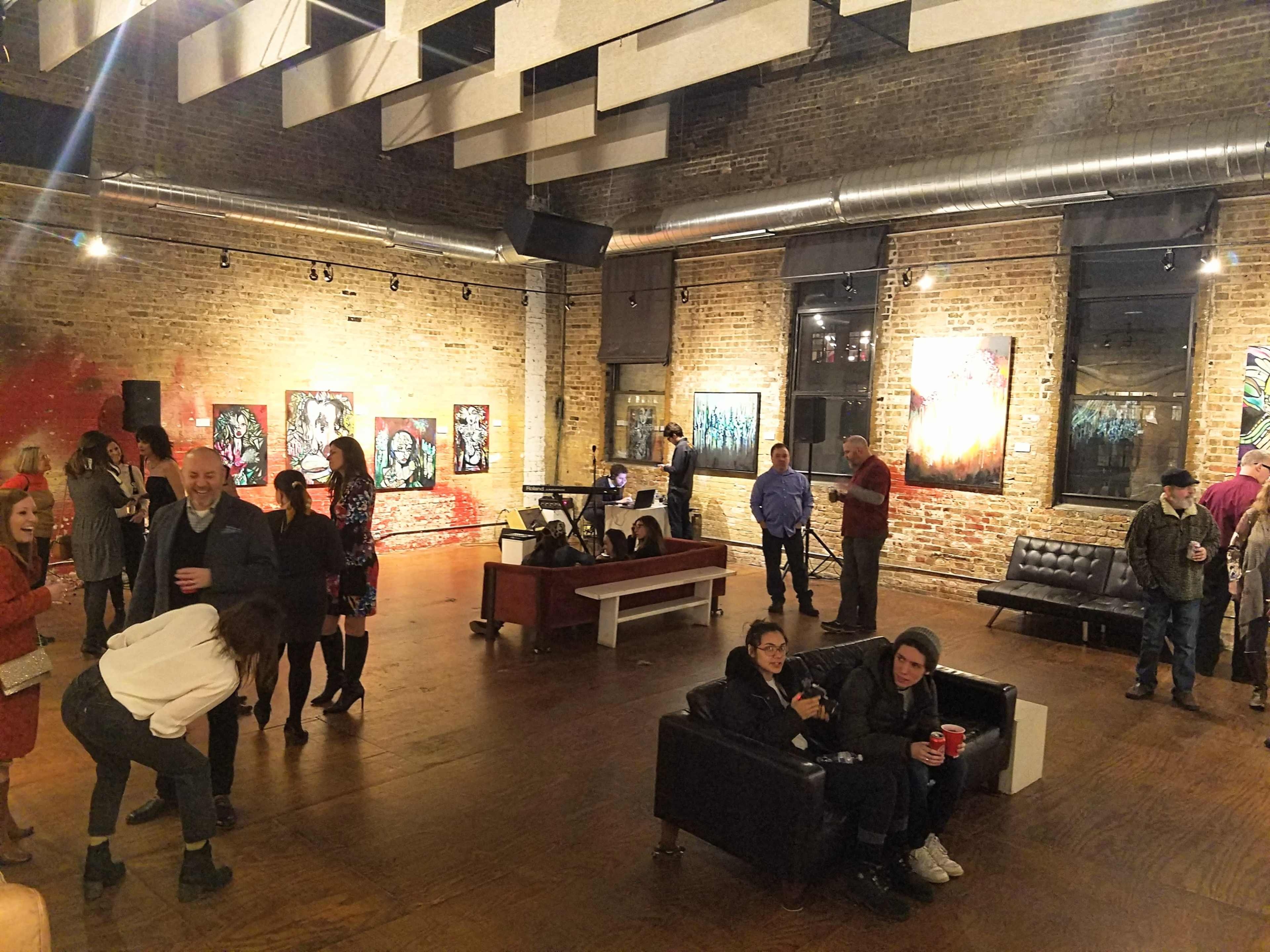 An art gallery event with attendees socializing amidst colorful artwork displayed on the brick walls.