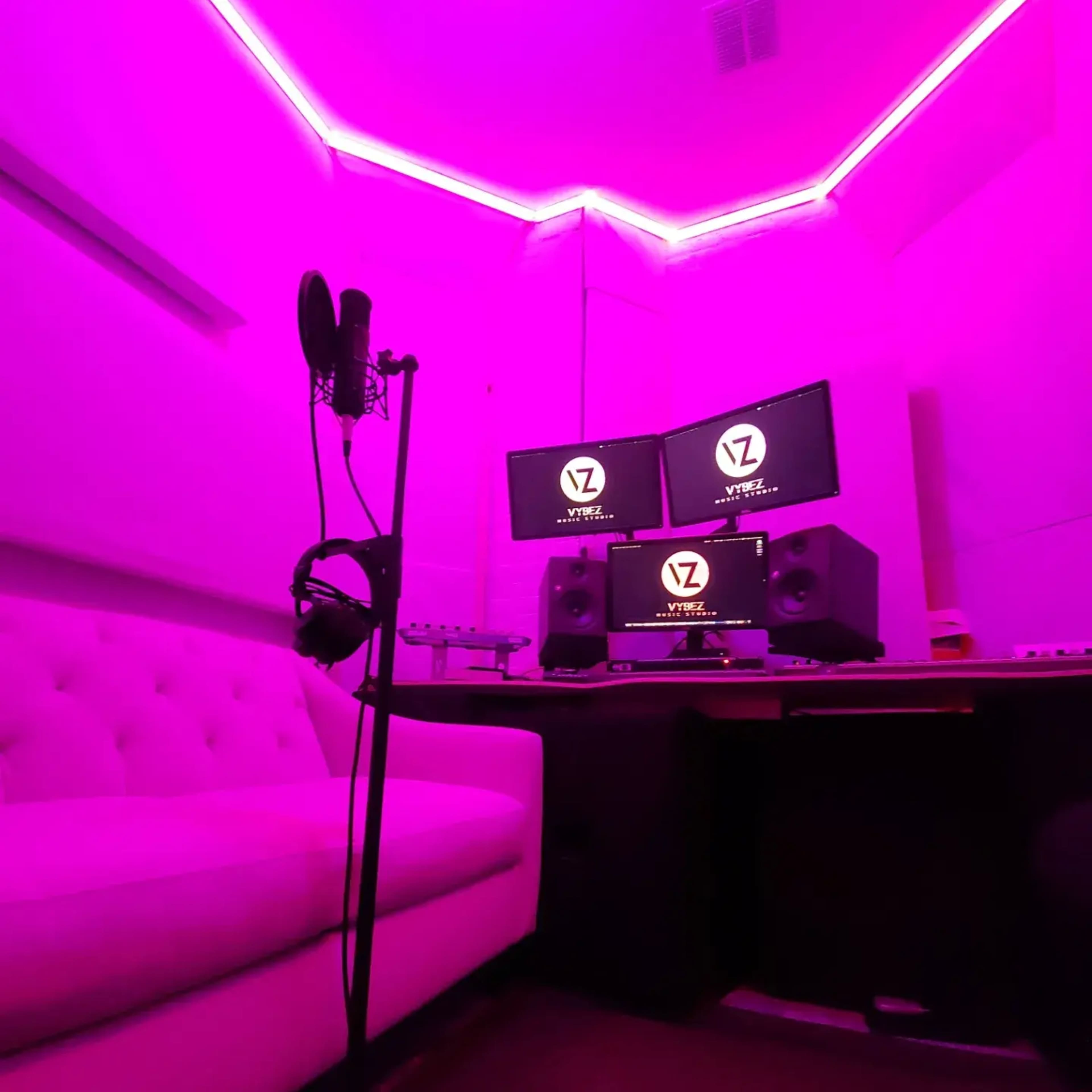 The image shows a music recording studio with pink lighting, a microphone stand, a computer setup with two monitors, and speakers.