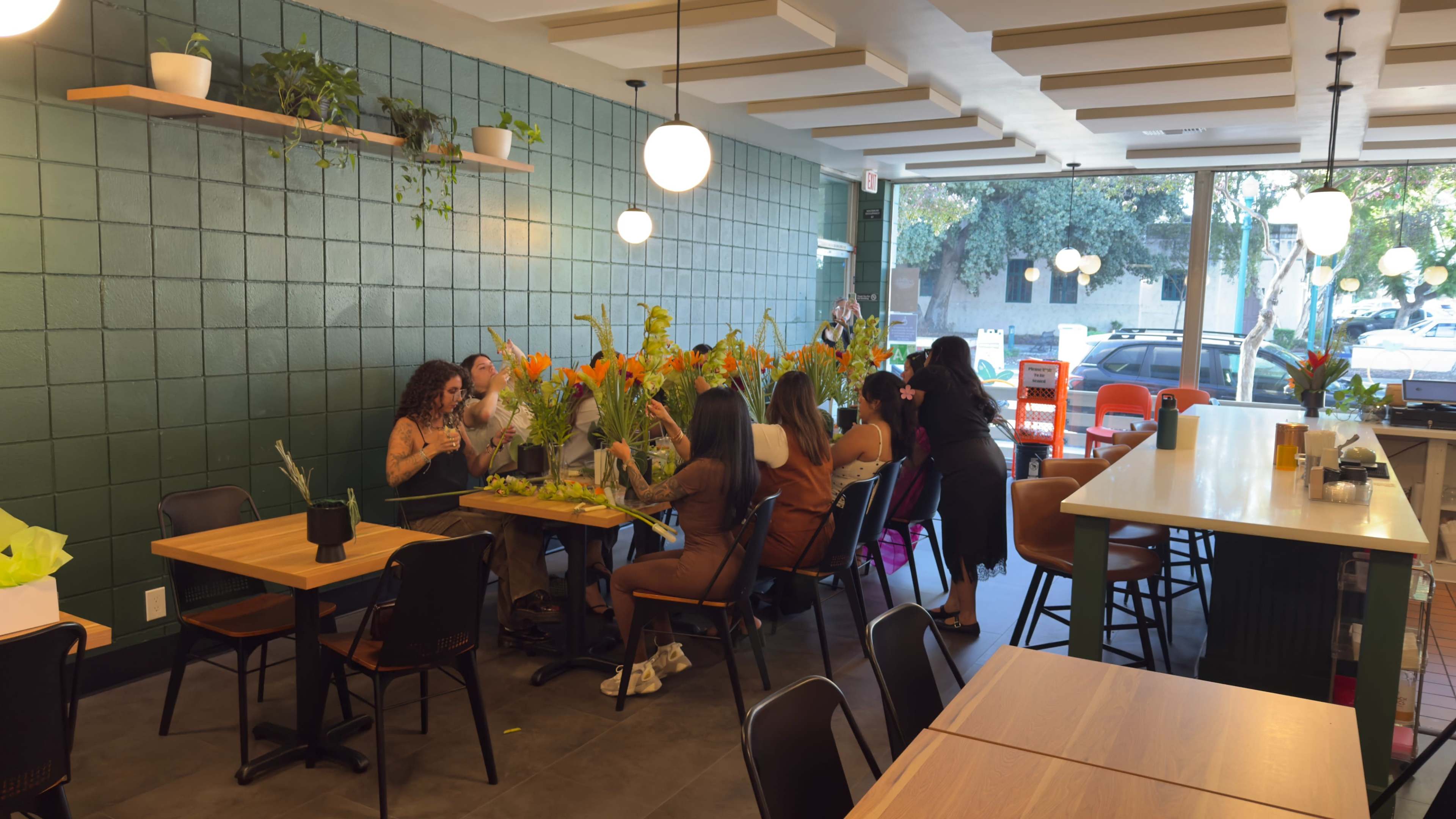 A group of people are gathered around a large table in a brightly lit café, arranging tropical flowers while others sit at adjacent tables.