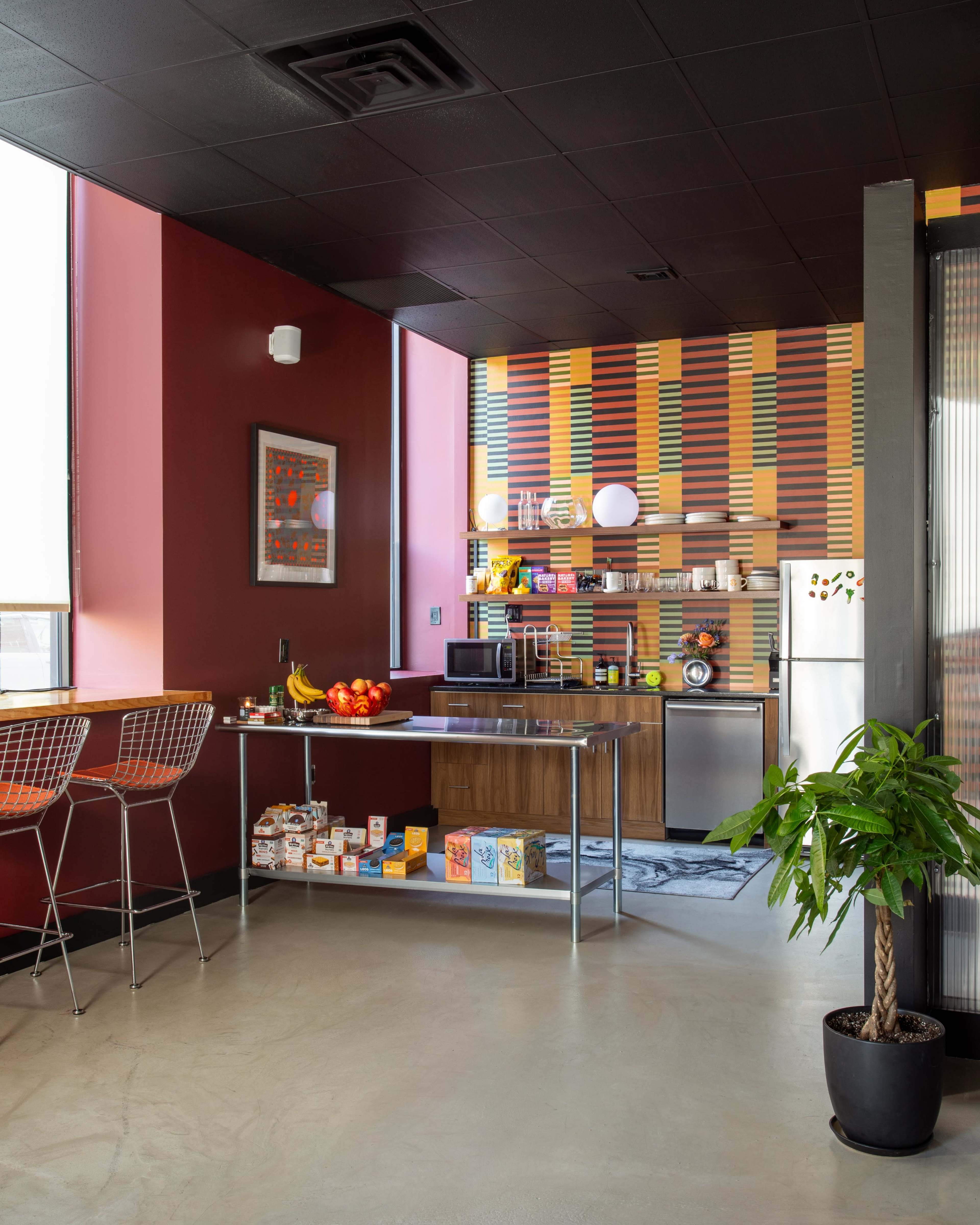 The image shows a modern kitchen area with a vibrant striped wall, a countertop with snacks, and seating with bar stools.