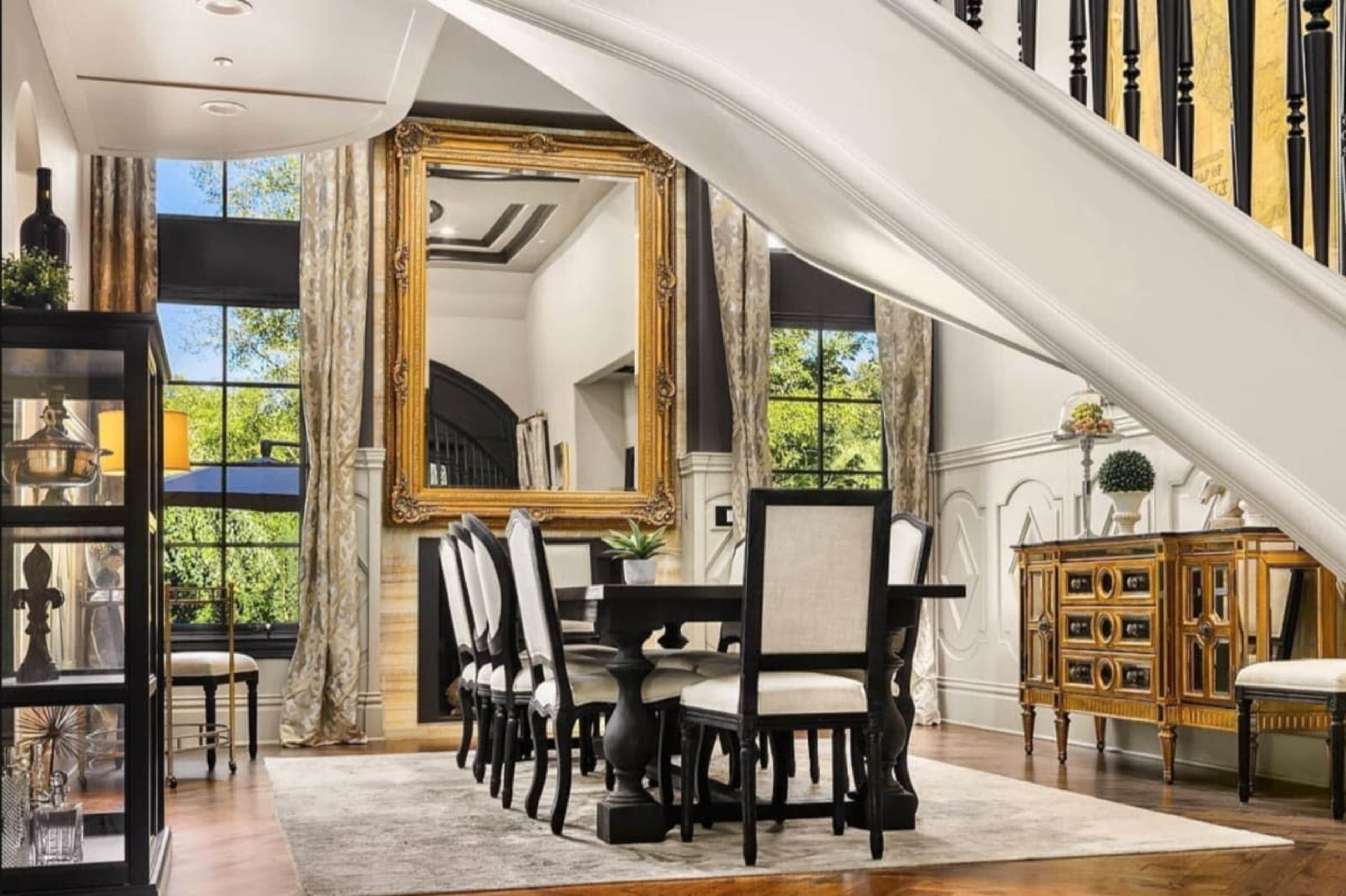 A dining area features a rectangular table surrounded by upholstered chairs, set beneath a large ornate mirror and a staircase.