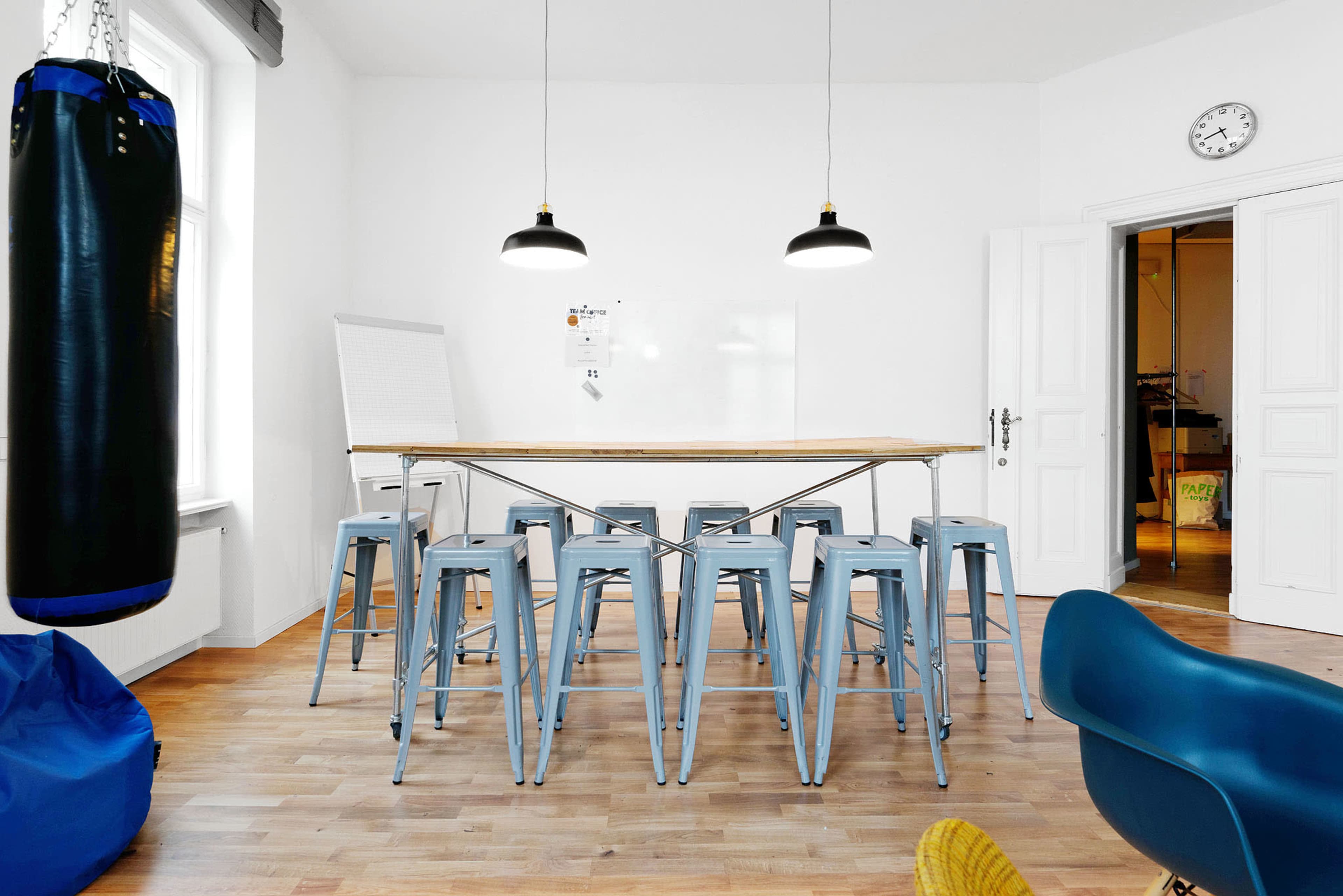 A spacious room with a long wooden table surrounded by metal stools, a punching bag in the corner, and double doors leading to another area.