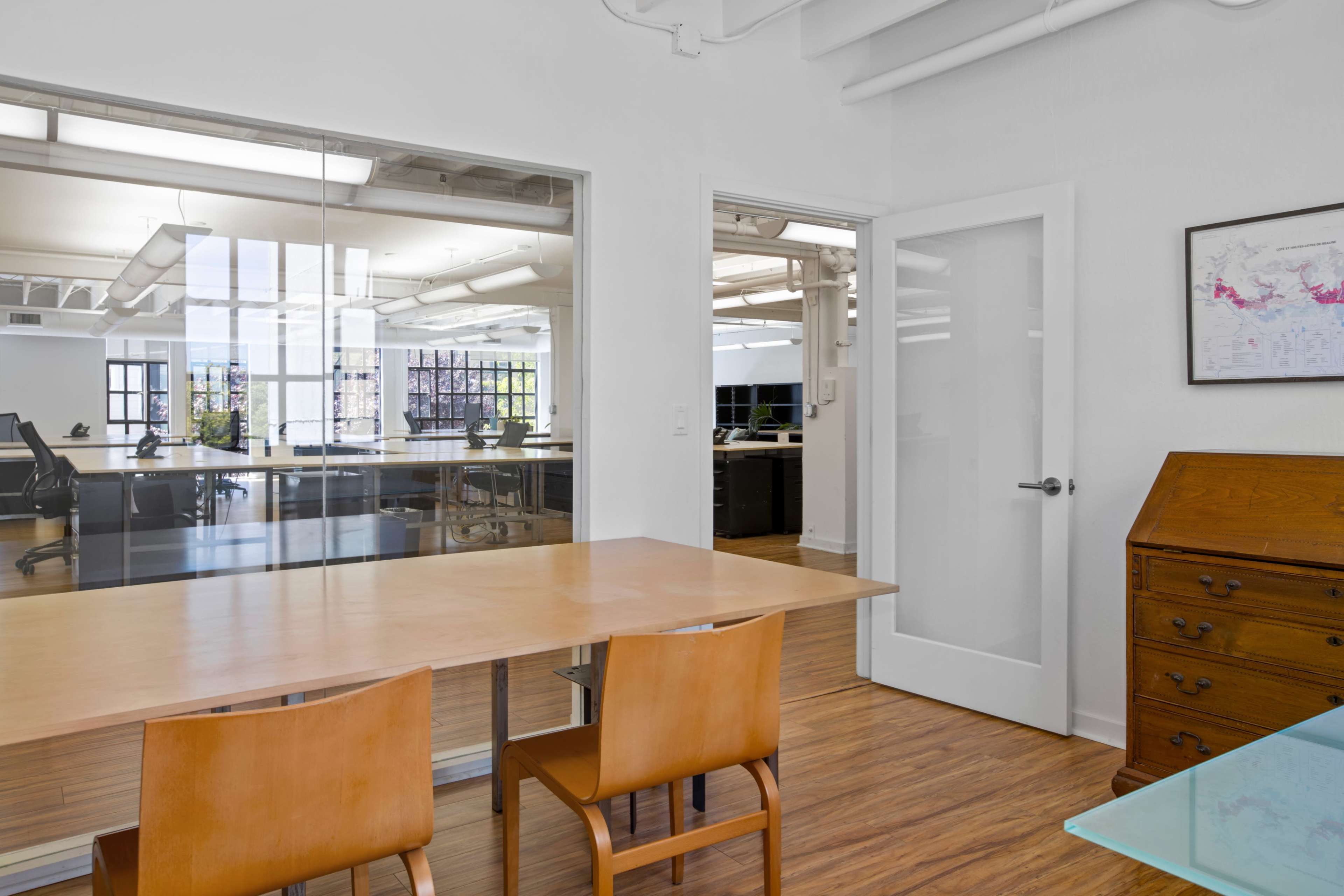 A bright office space features a wooden table with two chairs, surrounded by glass walls and doorways leading to additional work areas.