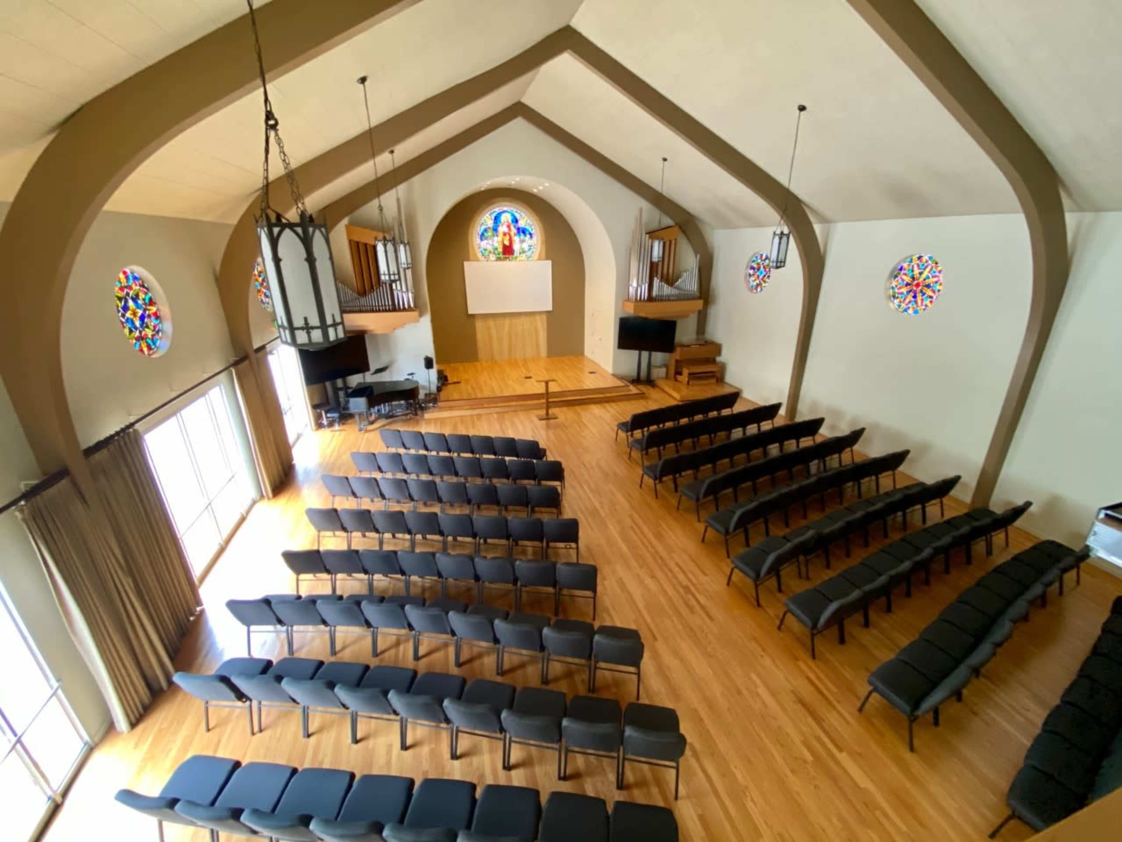An empty interior space of a church with rows of black chairs facing a stage and stained glass windows.