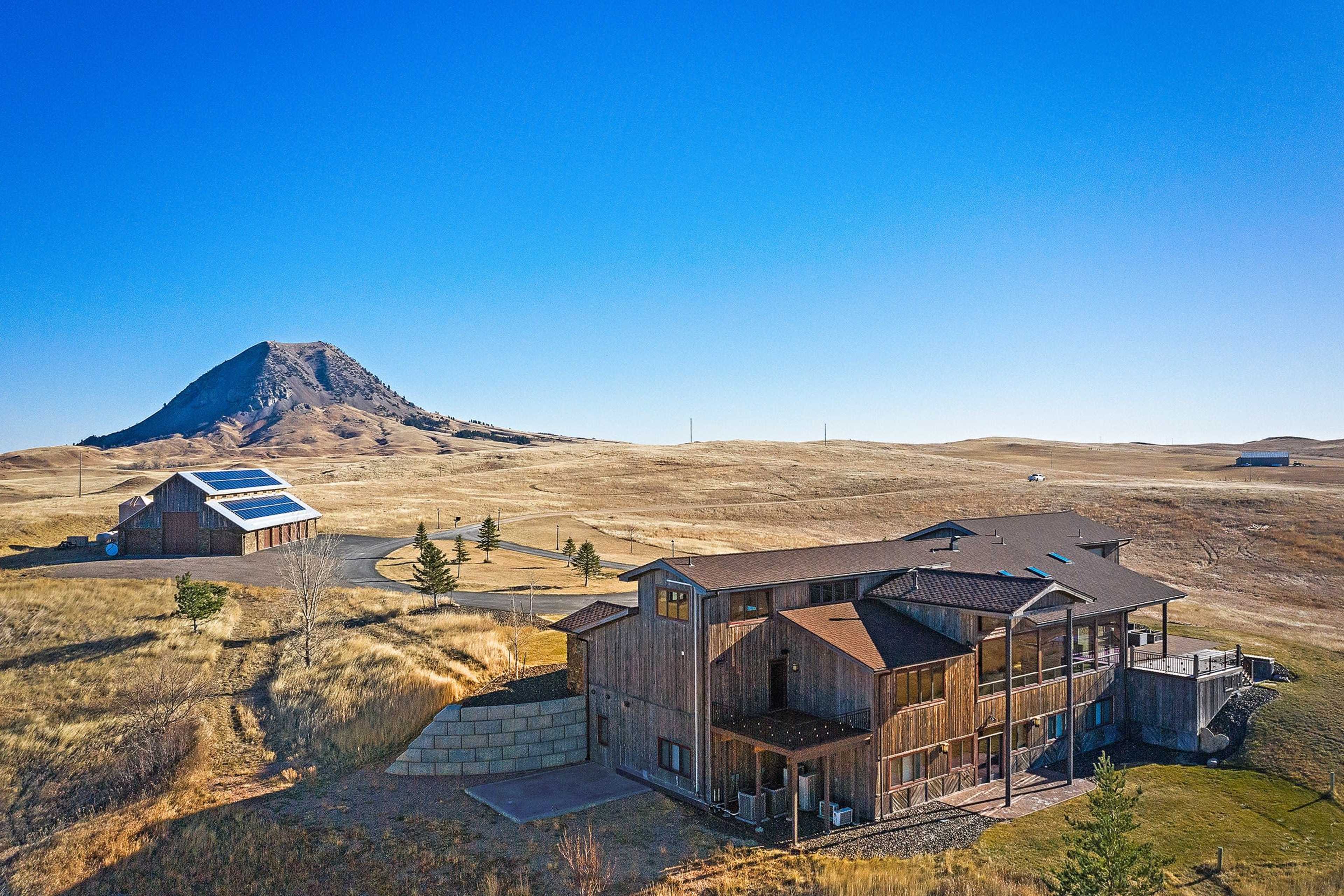 The image shows a large wooden house with a deck and a nearby structure featuring solar panels, set against a backdrop of rolling hills and a prominent, small mountain.