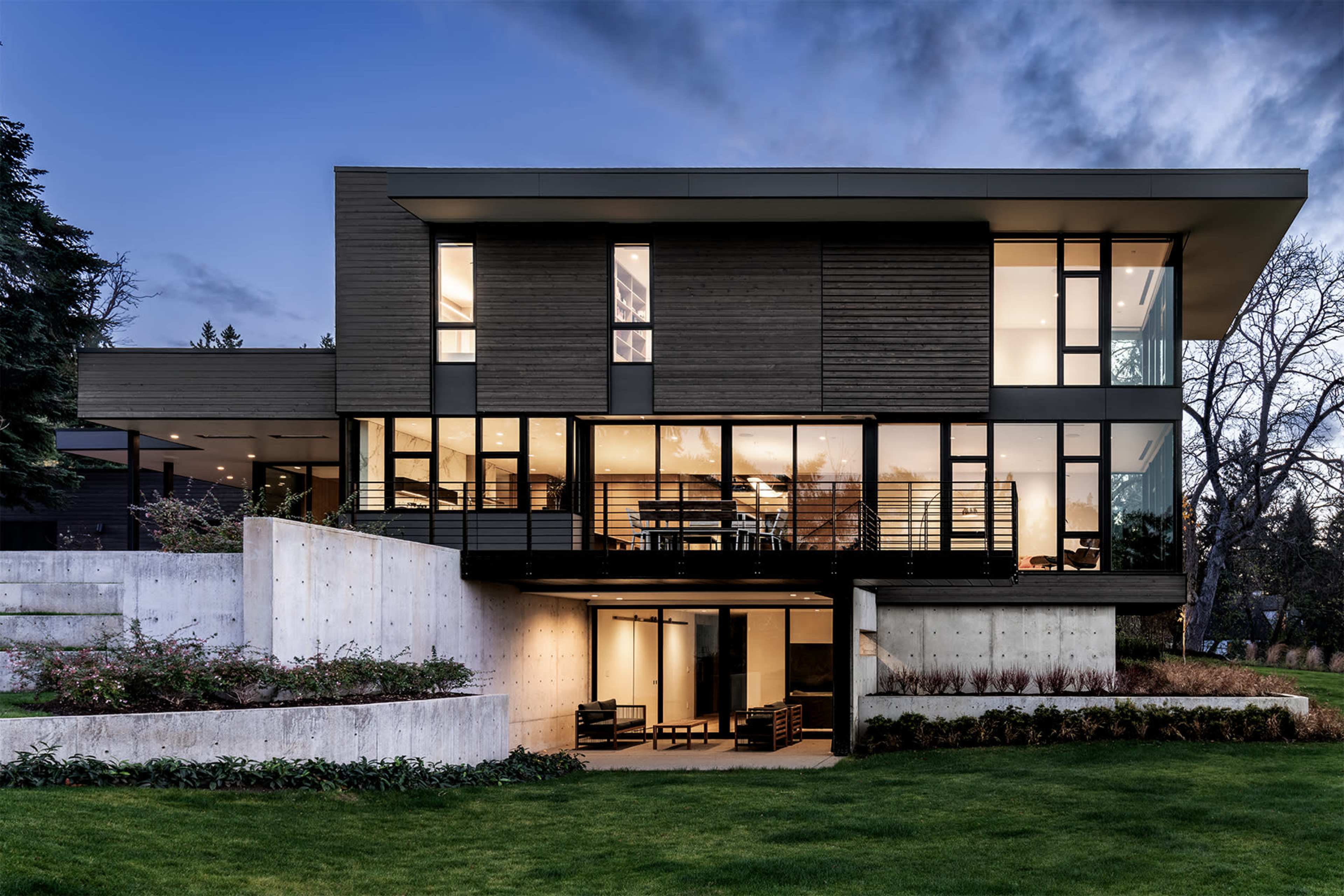 A modern two-story house with large windows and a mix of wood and concrete exterior sits on a grassy lawn.