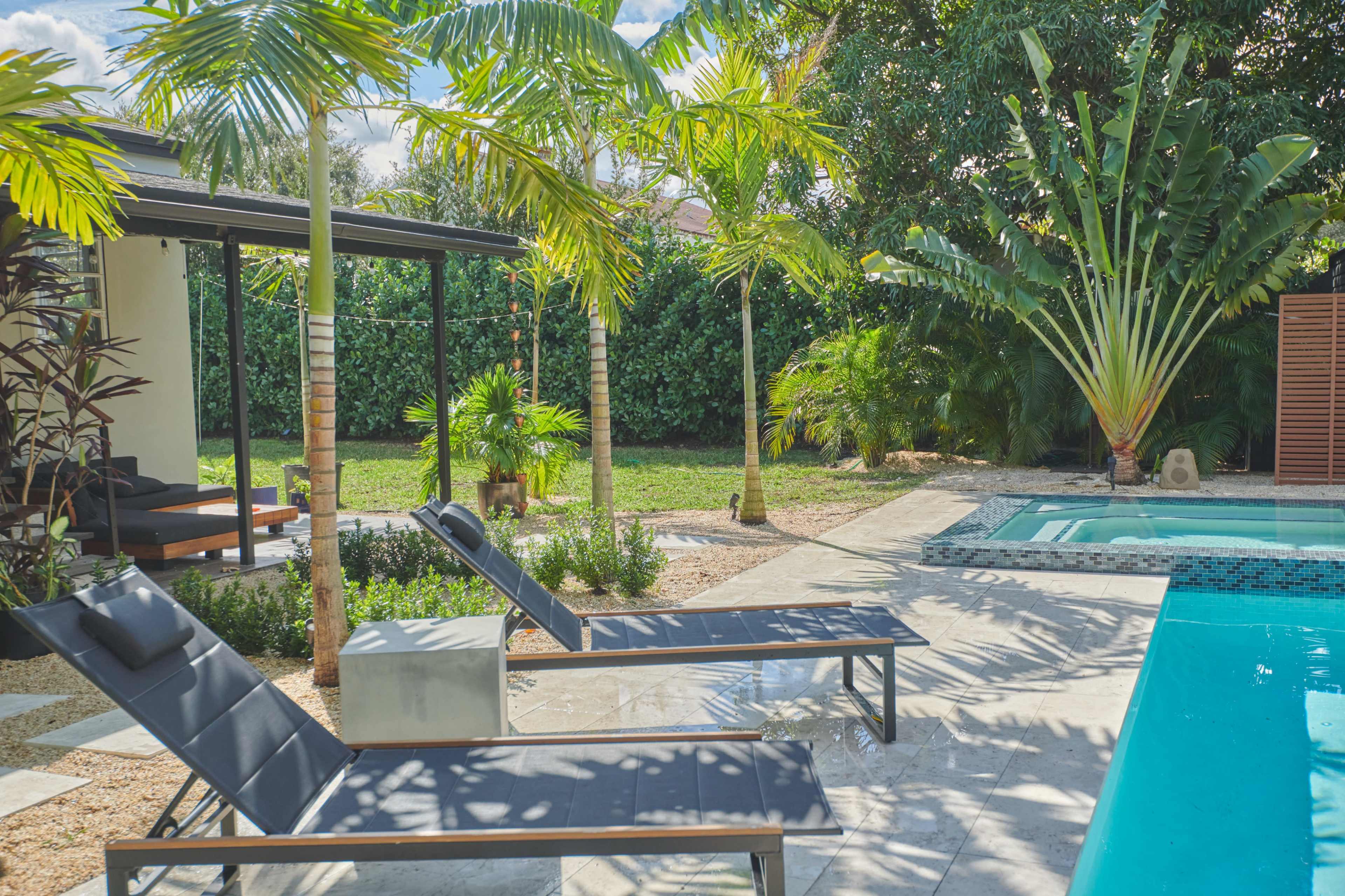 A serene outdoor space with two lounge chairs next to a swimming pool, surrounded by lush greenery and tropical plants.