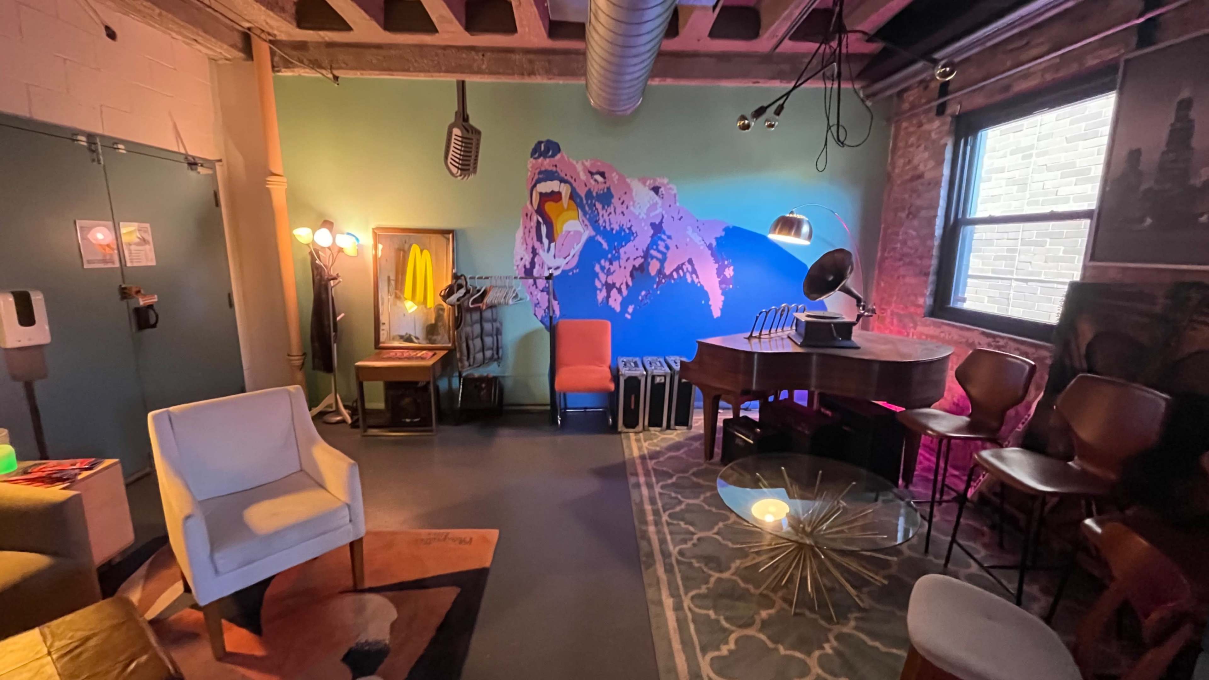The image shows a cozy room featuring a piano, colorful wall art of a dog, various seating options, and warm lighting fixtures.