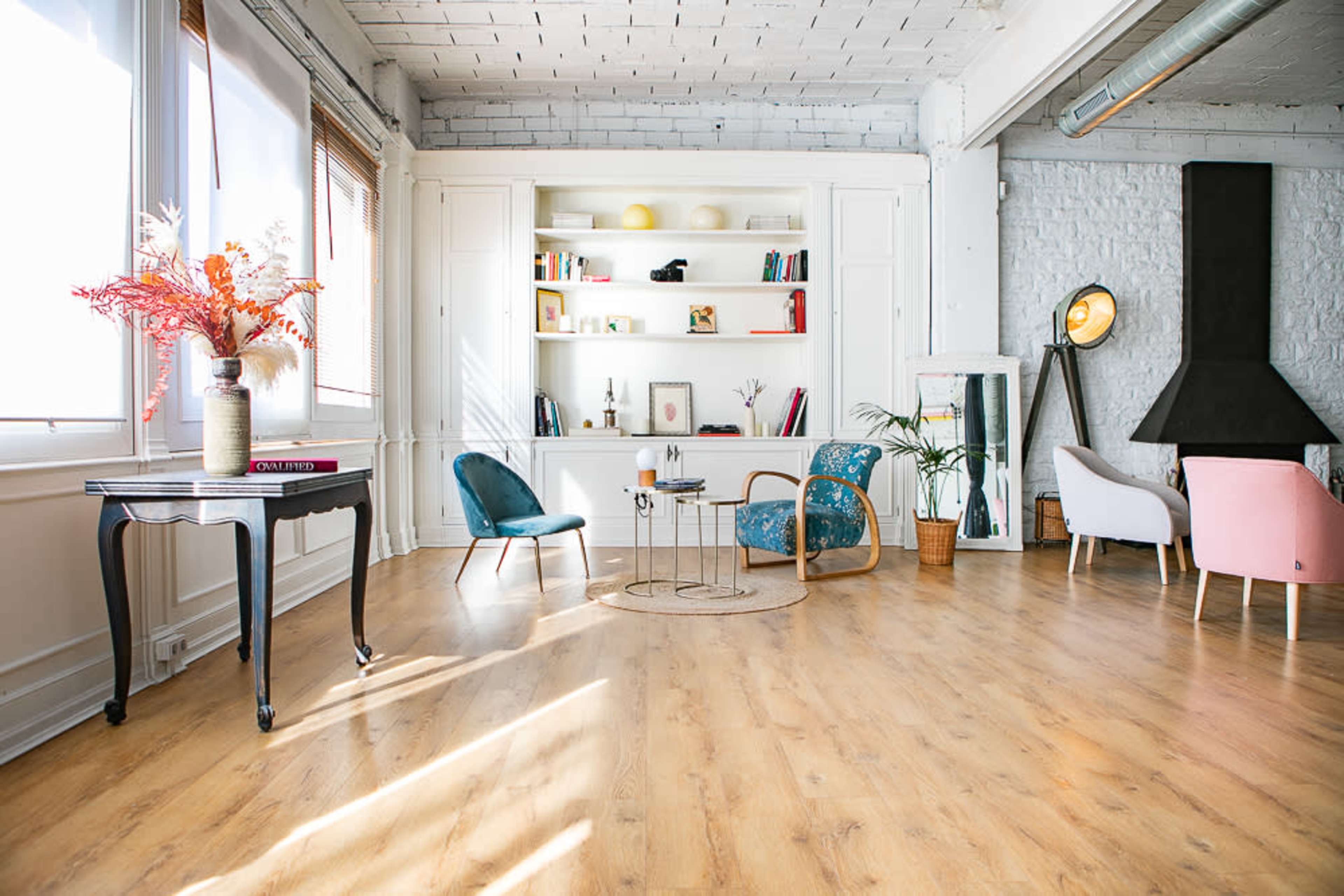 The image shows a bright, modern living space with a light wooden floor, a bookshelf filled with books and decorative items, and several stylish chairs arranged around a small table.